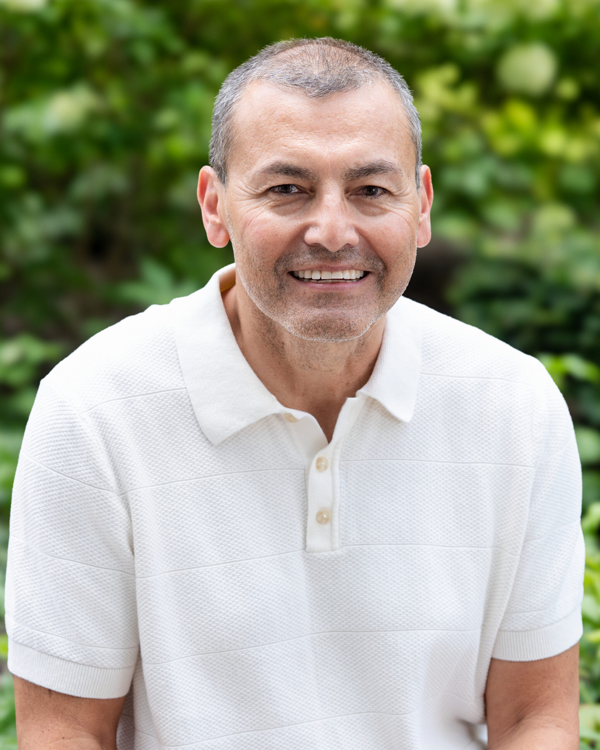 AWE-400-Troy - Alliance for Water Efficiency Smiling middle-aged man in a white polo shirt outdoors, with green blurred foliage in the background.
