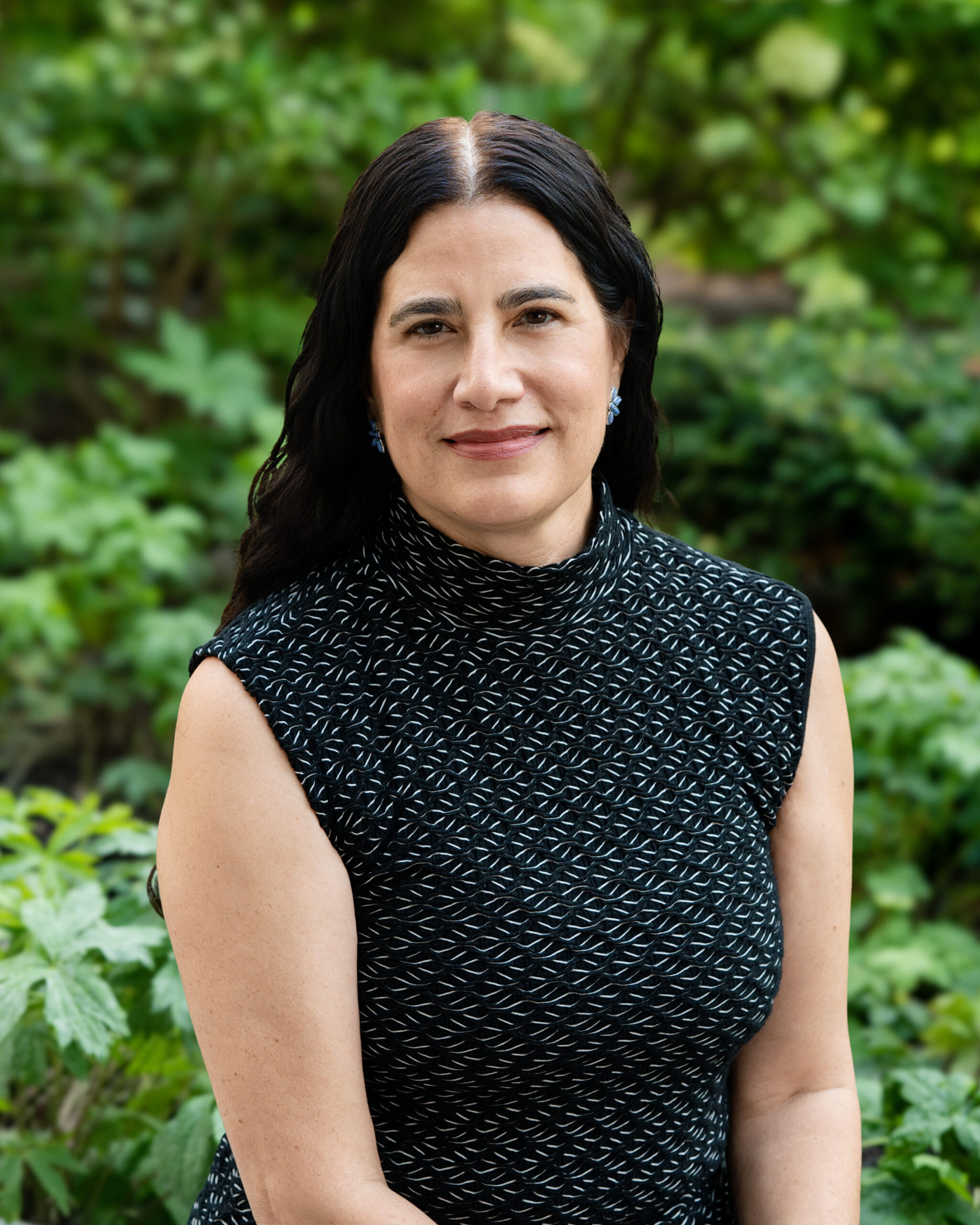 AWE-184-Christine - Alliance for Water Efficiency Portrait of a woman with dark hair wearing a black patterned sleeveless top, smiling with a lush green garden behind her.