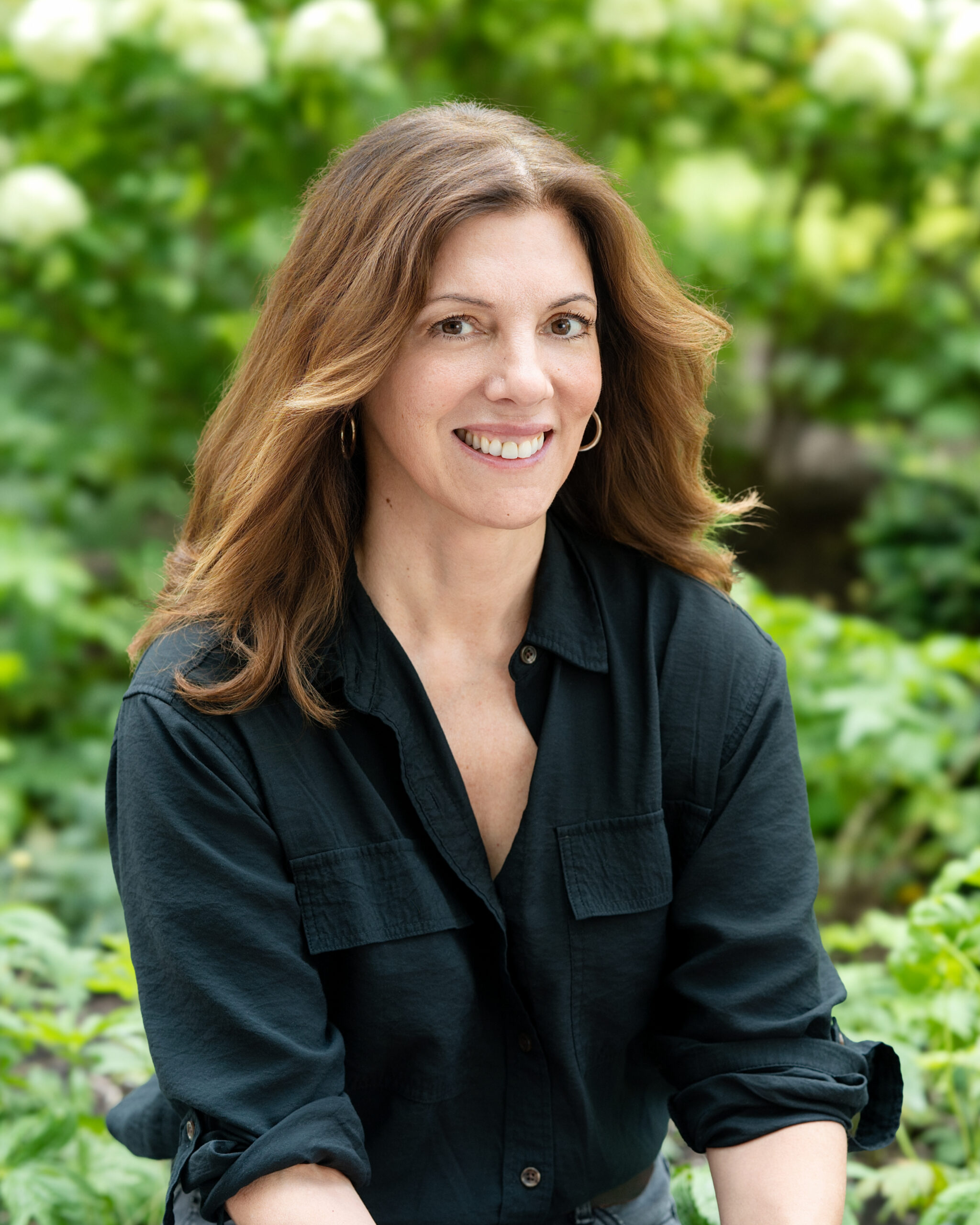 AWE-417-Paula - Alliance for Water Efficiency Portrait of a smiling woman with long brown hair wearing a dark shirt, outdoors with greenery in the background.