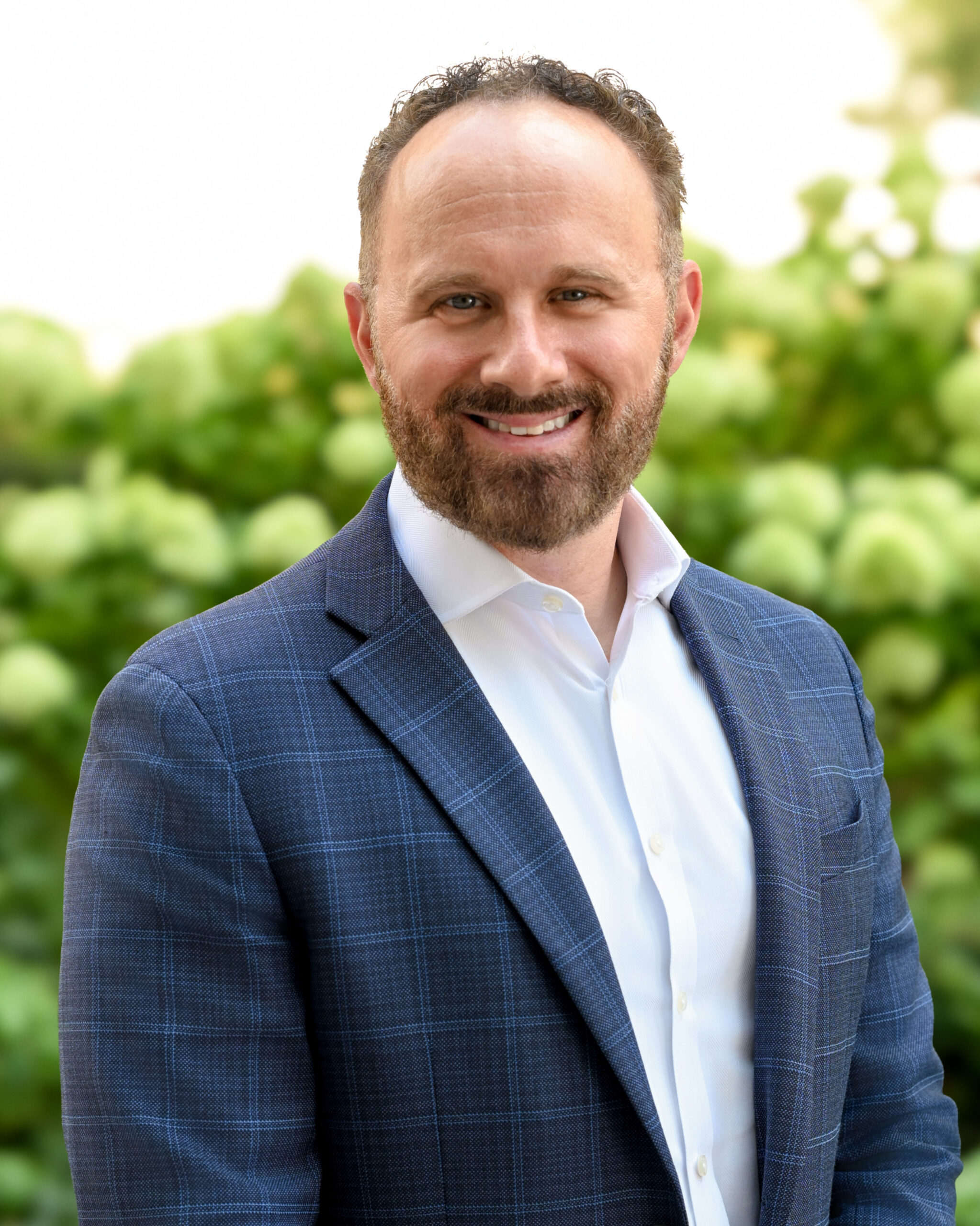 AWE138-Dain - Alliance for Water Efficiency Smiling man in a blue plaid blazer and white shirt, outdoors with green foliage in the background.