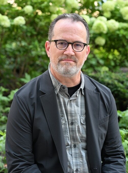 AWE246 - Alliance for Water Efficiency Portrait of a middle-aged man with glasses, grey beard, wearing a dark blazer over a checkered shirt, outdoors among greenery.