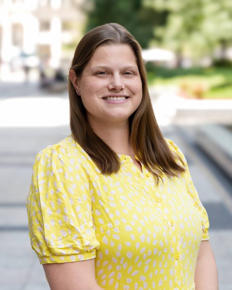 Jill-Greiner-updatedImg - Alliance for Water Efficiency Smiling woman with brown hair in a yellow patterned blouse outdoors, urban park background behind her.