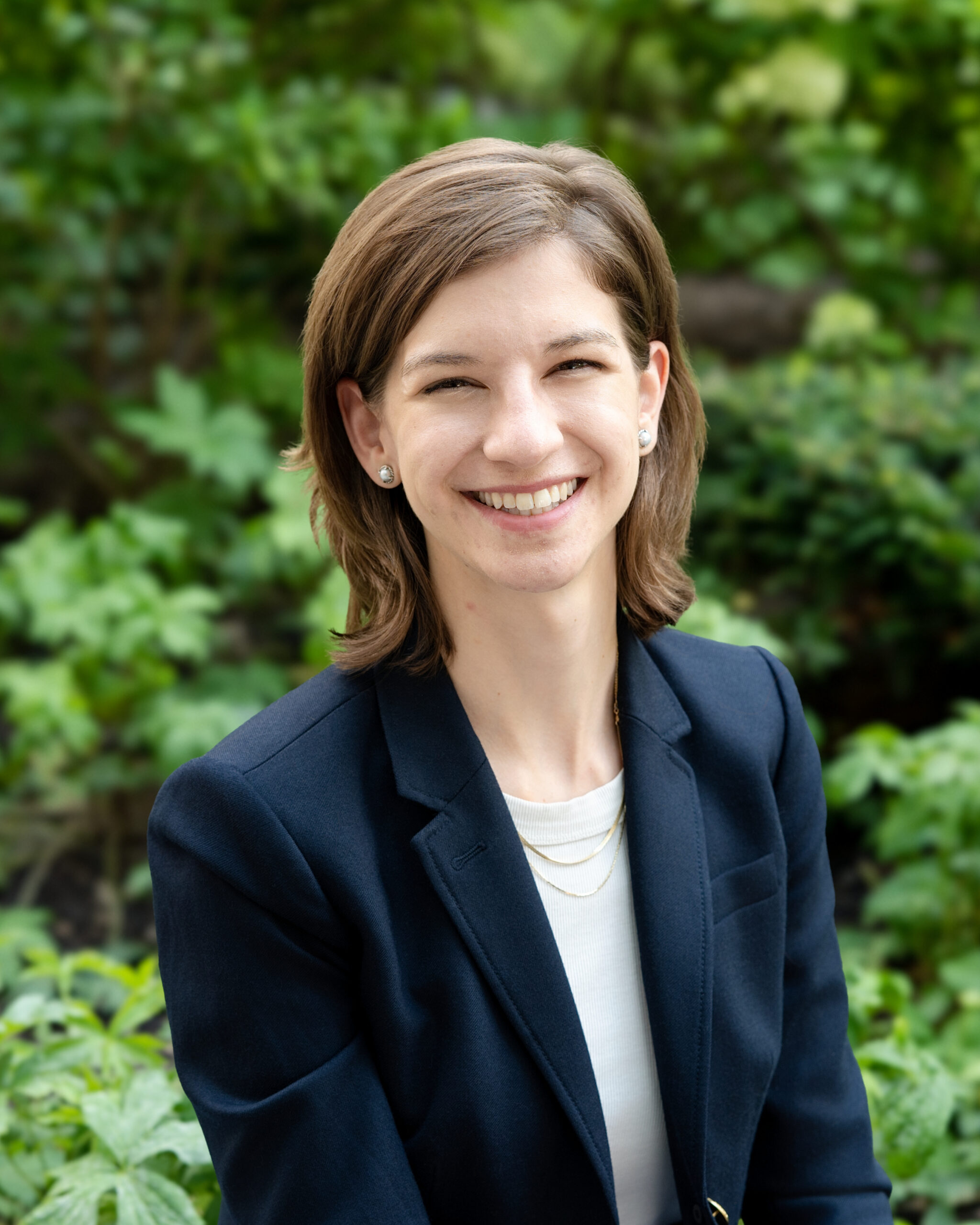 AWE-260-Devin - Alliance for Water Efficiency Portrait of a smiling woman with short brown hair, wearing a navy blazer, outdoors with green foliage behind.══════════════════════=