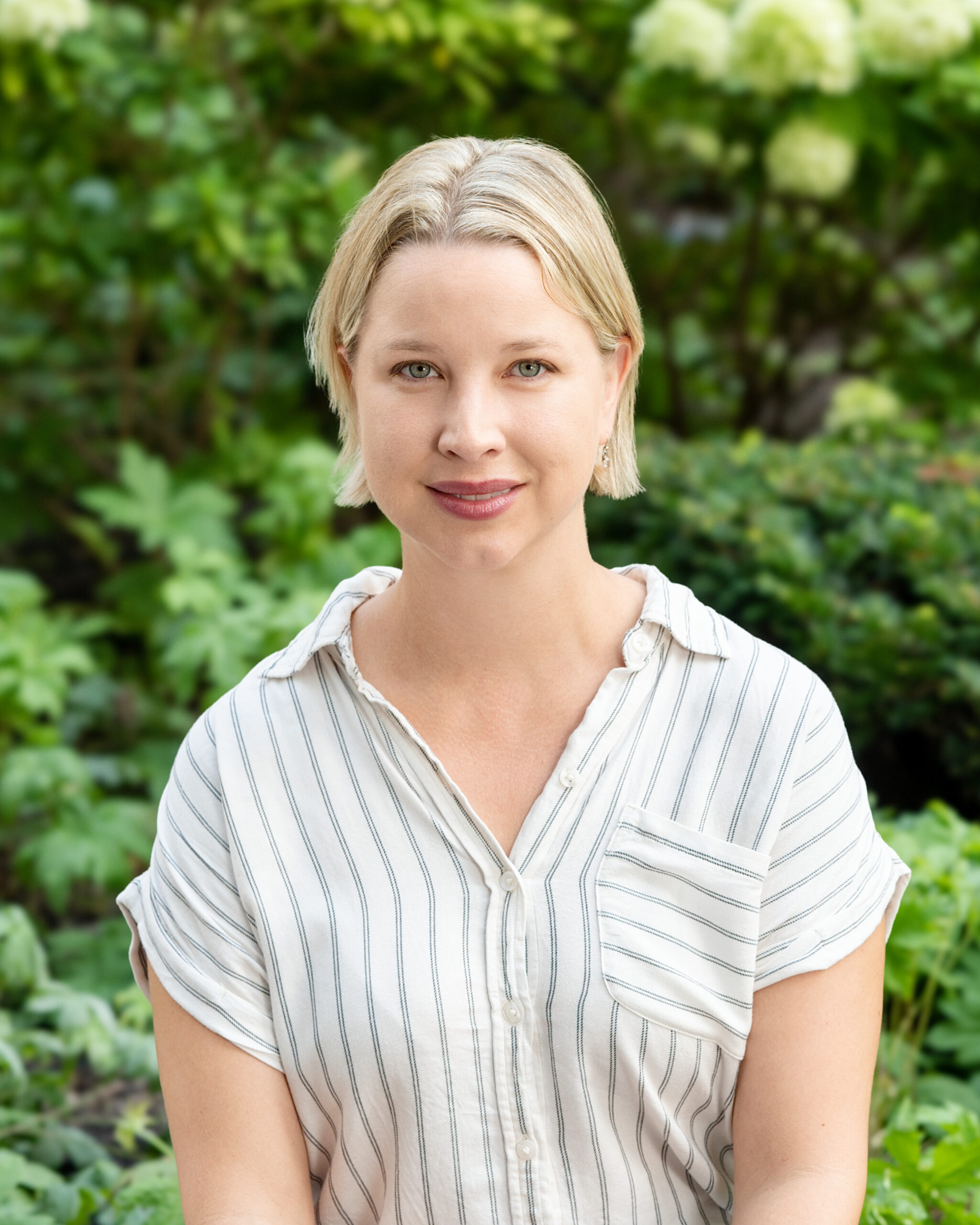 AWE-Sophie1 - Alliance for Water Efficiency Portrait of a woman with short blonde hair outdoors, wearing a white striped shirt, green foliage in the background