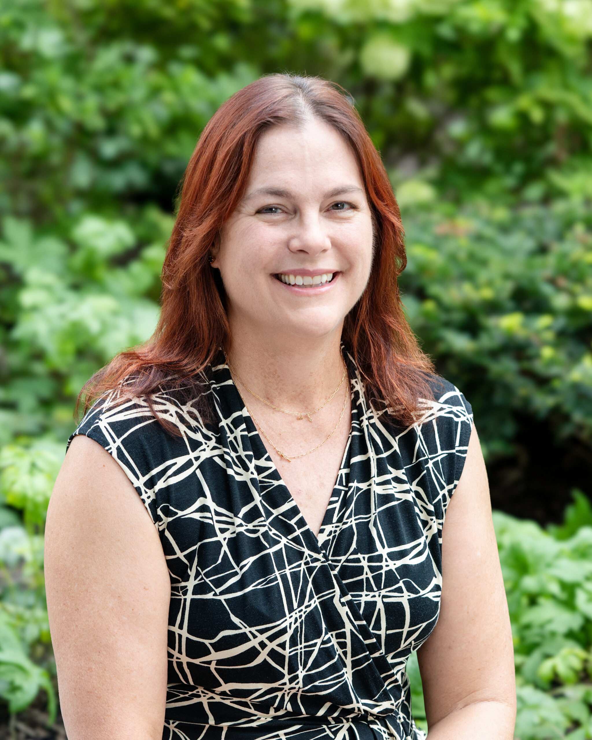 AWE-Joan 2 - Alliance for Water Efficiency Smiling woman with reddish hair outdoors, green foliage in the background.