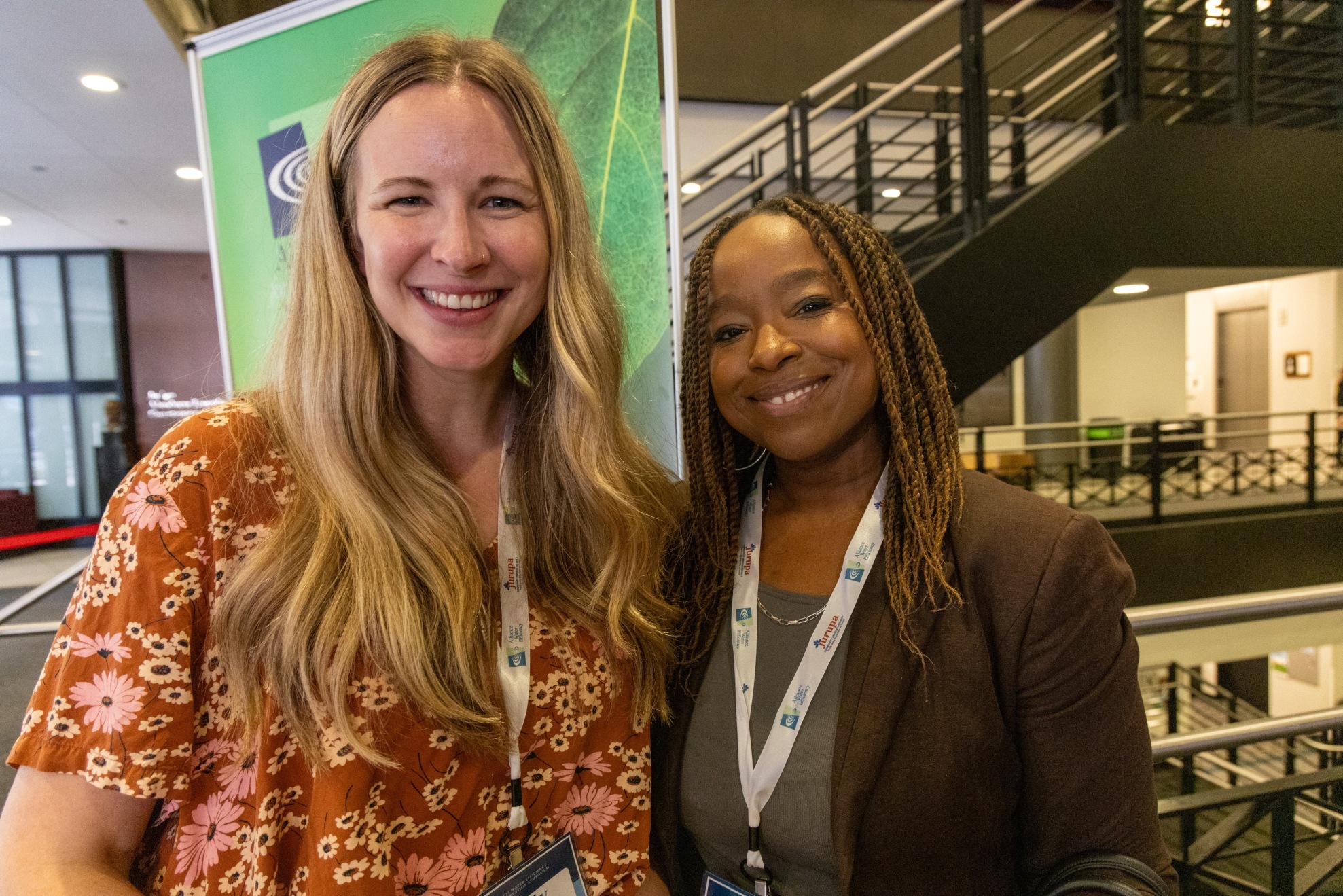 Two smiling women at a conference, wearing lanyards, posing together indoors with a green banner behind them.