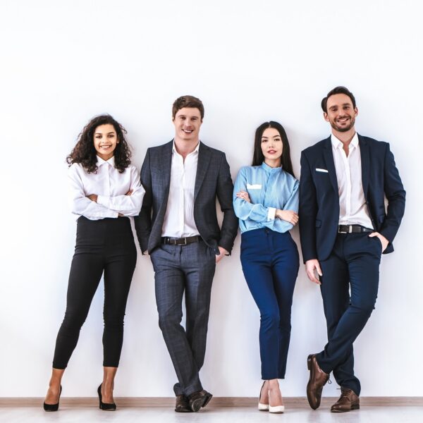 Five diverse professionals in business attire stand against a white wall, smiling for a team photo.