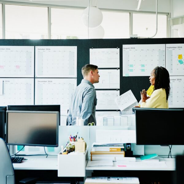 Two coworkers standing and talking in front of a wall covered with calendars and notes in an office.