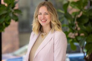 Smiling woman with blonde hair wearing a beige sweater and light pink blazer, outdoors among green plants.