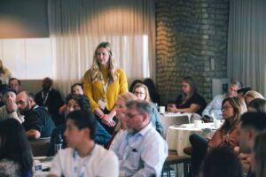 Woman in mustard sweater standing and speaking at a conference, with attendees seated around tables listening nearby