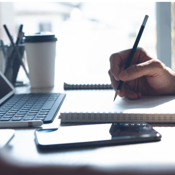 Close-up of a hand writing with a pencil at a desk, with a laptop and notebook nearby.