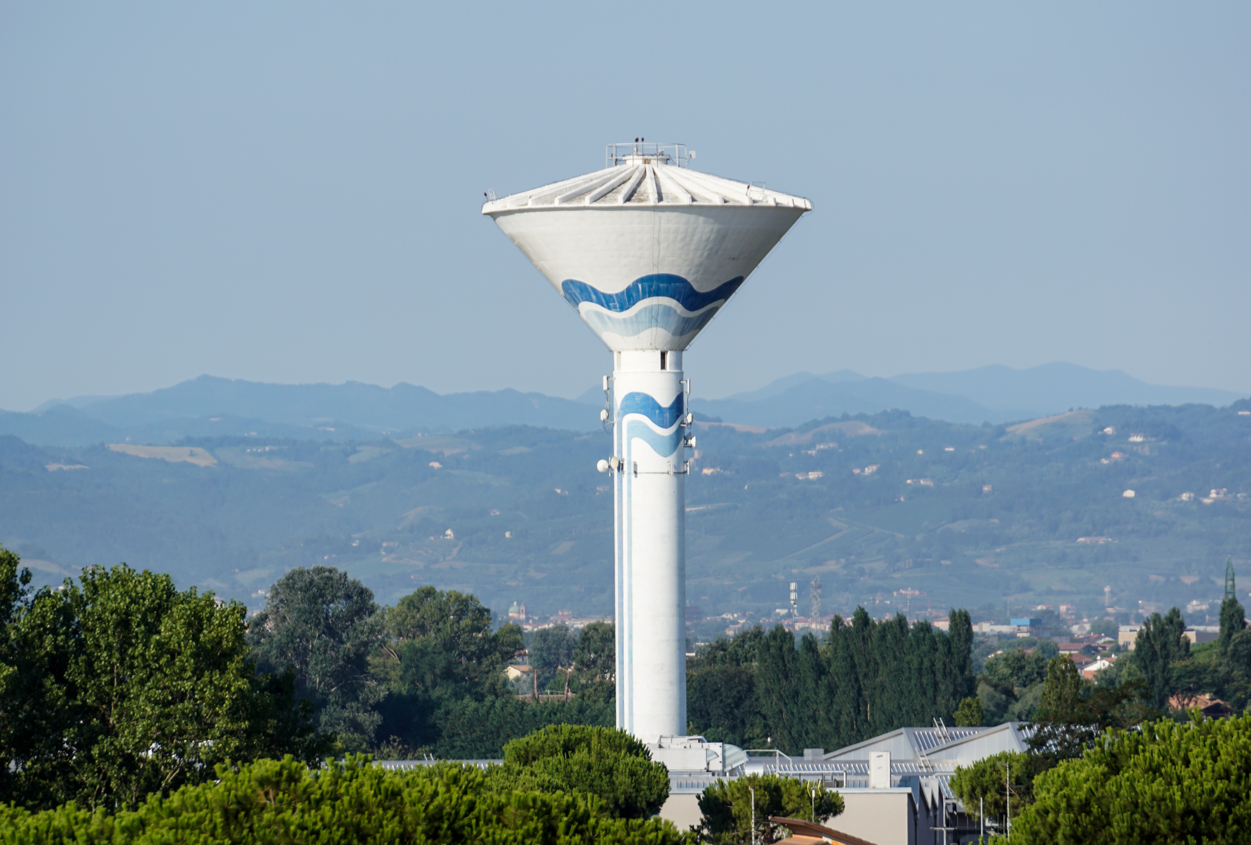 White water tower with a blue wave mural, set against a clear blue sky and distant hills.