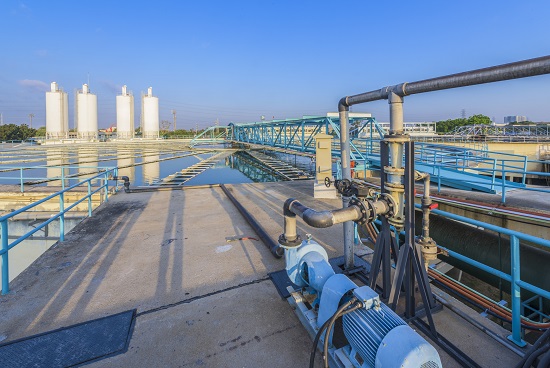 Industrial water treatment site with a large blue pump and network of pipes on a concrete platform, blue railings, and a walkway extending toward white tanks in the distance under a clear blue sky