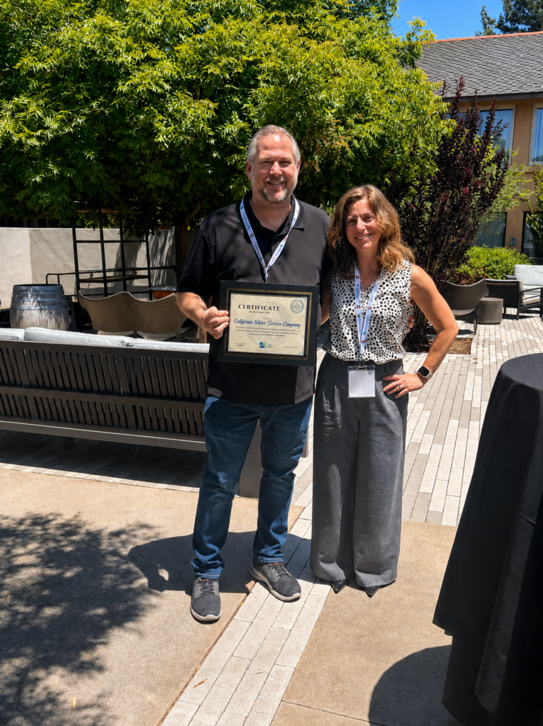 Two adults stand outdoors, smiling, while holding a framed certificate after an award event.
