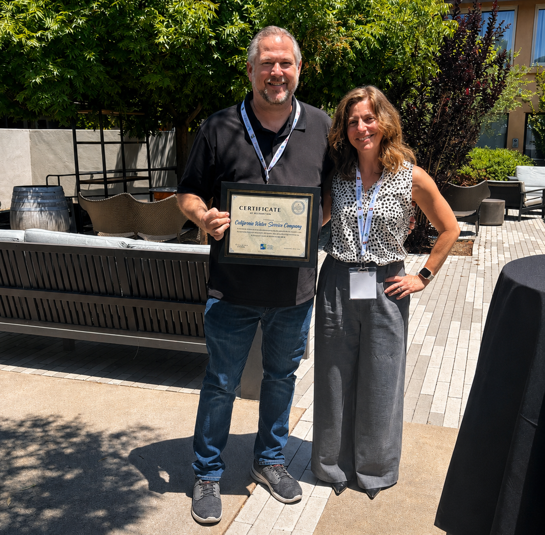 Two adults stand outdoors, smiling, while holding a framed certificate after an award event.