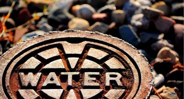 Close-up of a circular metal water manhole cover with the word WATER and a gear-like pattern on a rocky ground
