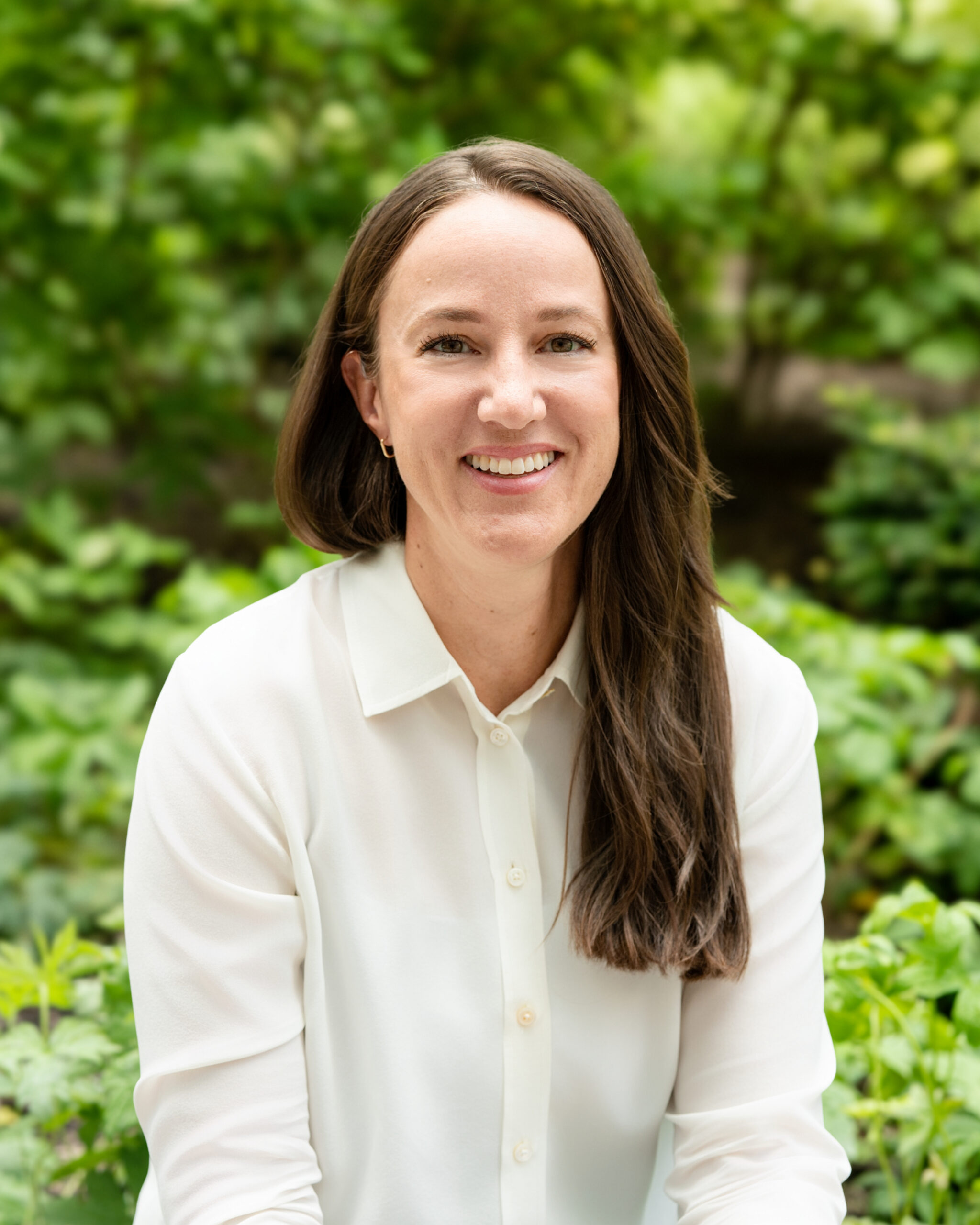 AWE-362-Meryl - Alliance for Water Efficiency Smiling woman with long brown hair in a white blouse, outdoors with lush green foliage in the background.