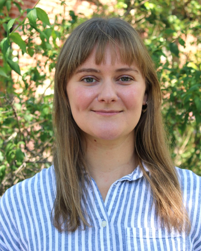 Smiling woman with light brown hair and bangs outdoors, wearing a blue-and-white striped shirt with greenery in the background.