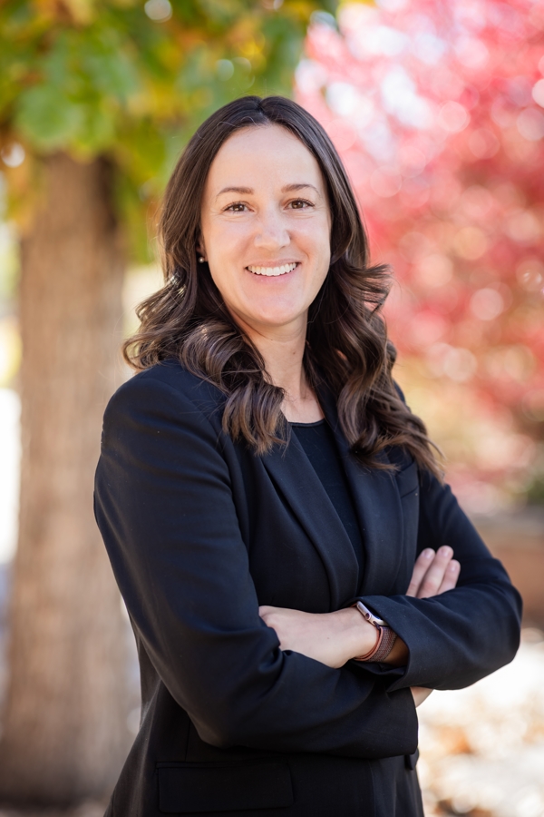 Meryl Headshot 1 - Alliance for Water Efficiency Professional woman with dark hair in a black blazer, arms crossed, smiling outdoors with autumn-colored foliage behind her.