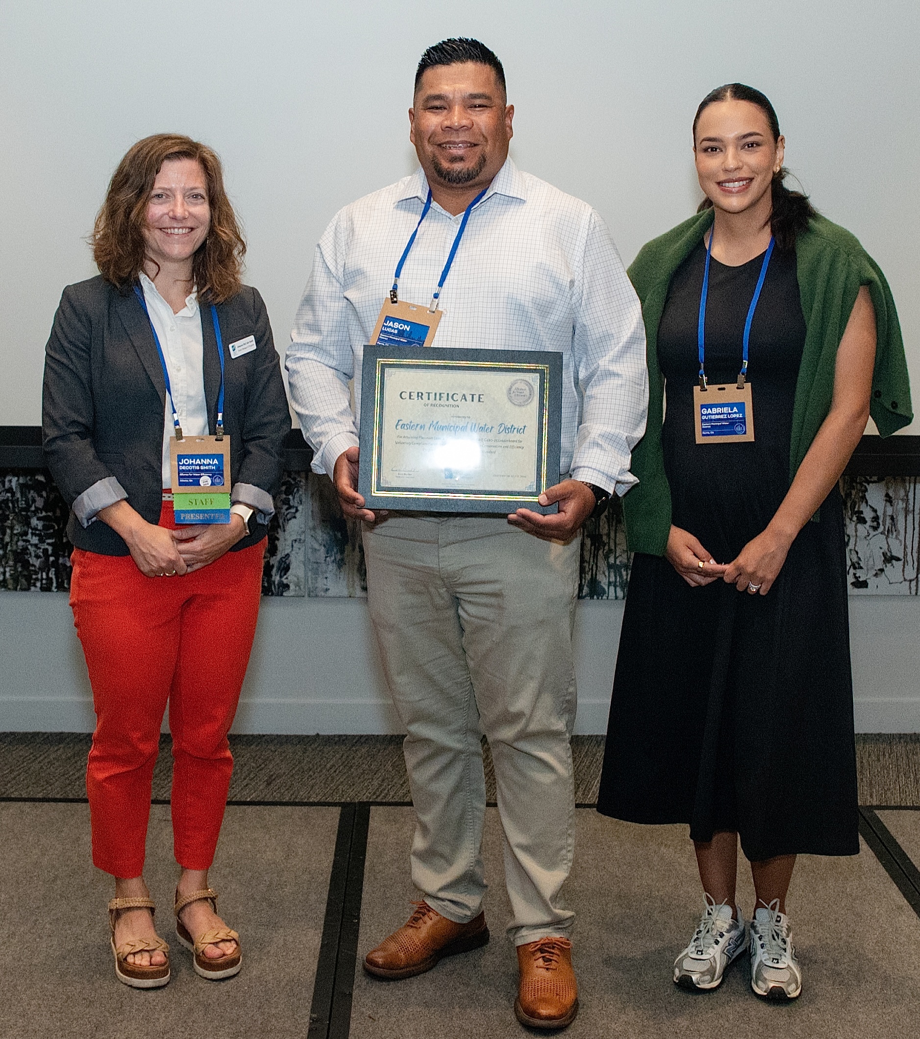 Three people stand side by side; the man in the center holds a framed certificate while the two women flank him, smiling warmly.