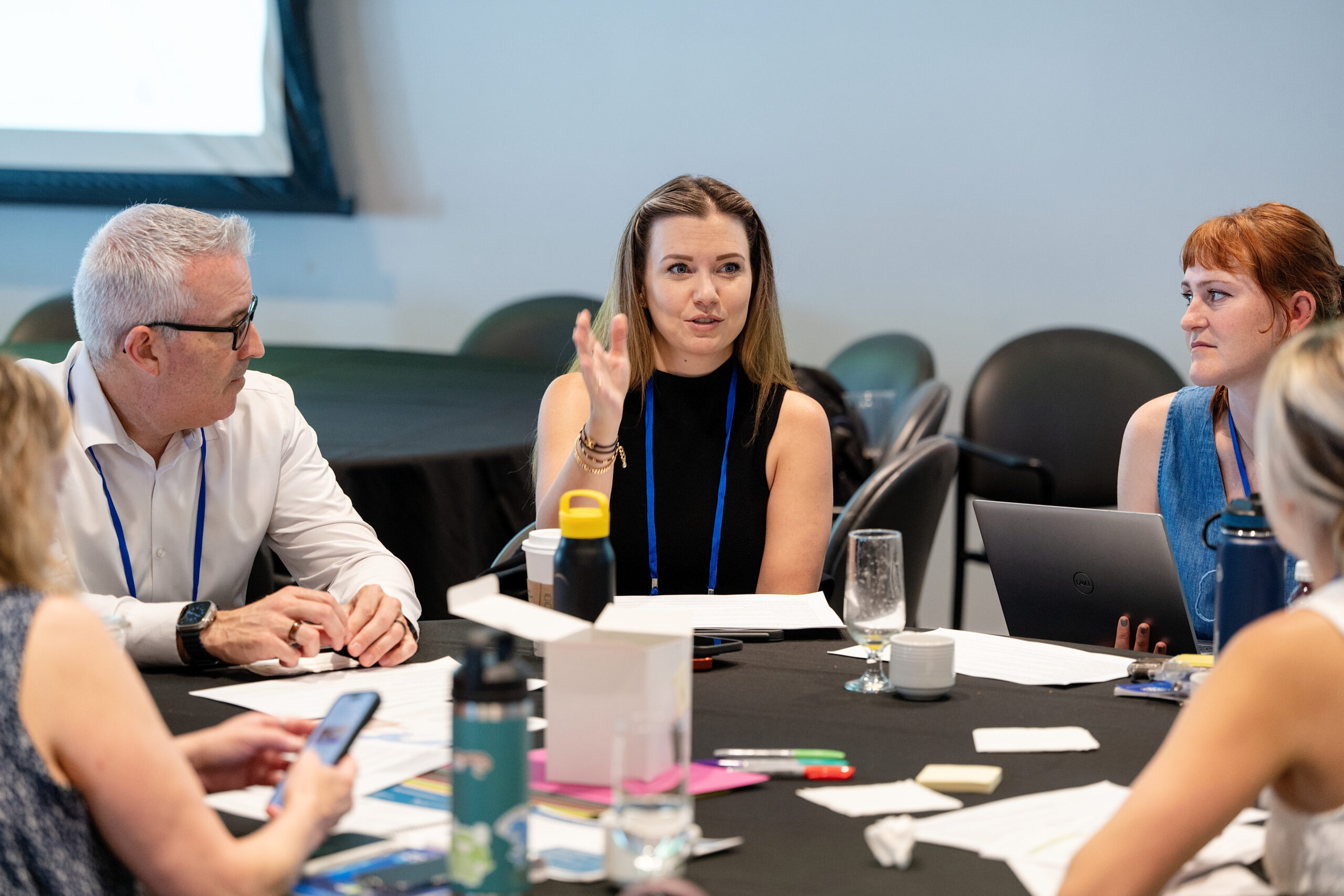 Group of colleagues at a meeting around a table, the woman in center speaking and gesturing with her hand.