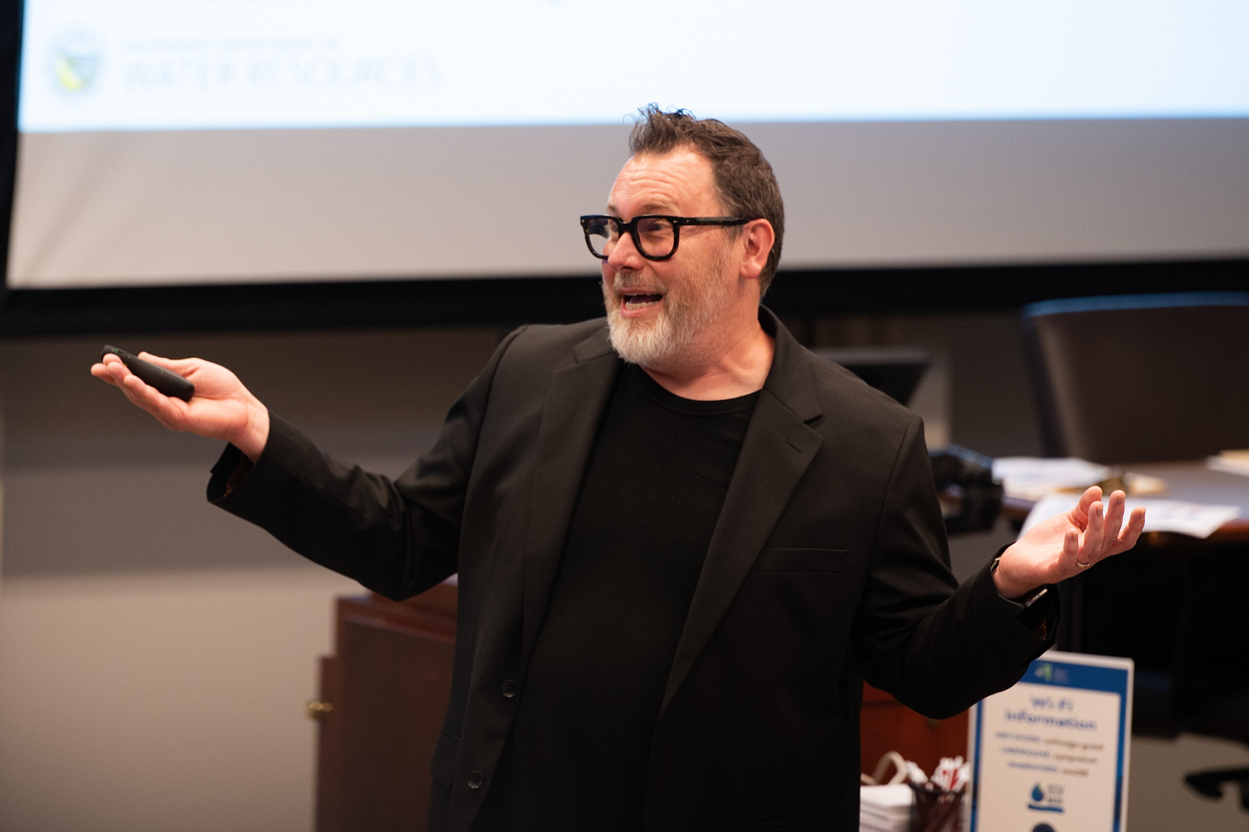 Bearded man with black-framed glasses in a dark blazer speaking and gesturing during a presentation in a conference room.