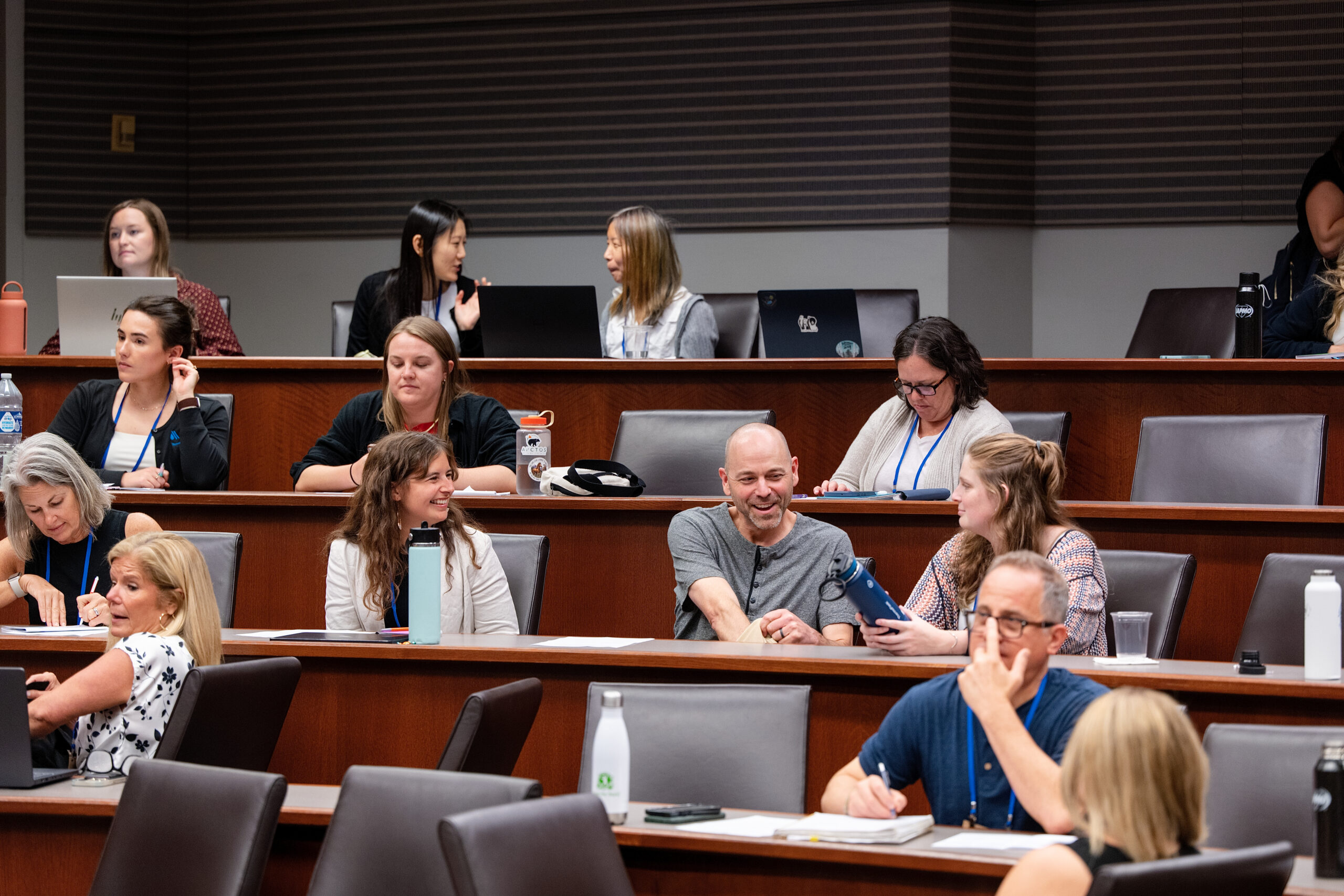 Diverse group of adults seated in a tiered classroom, some with laptops and notebooks, some talking in the back row while others work.