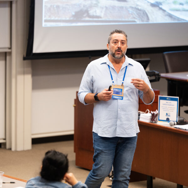 Male presenter in a light blue shirt with a blue lanyard and badge speaking to an audience in a seminar room, projector screen behind him.