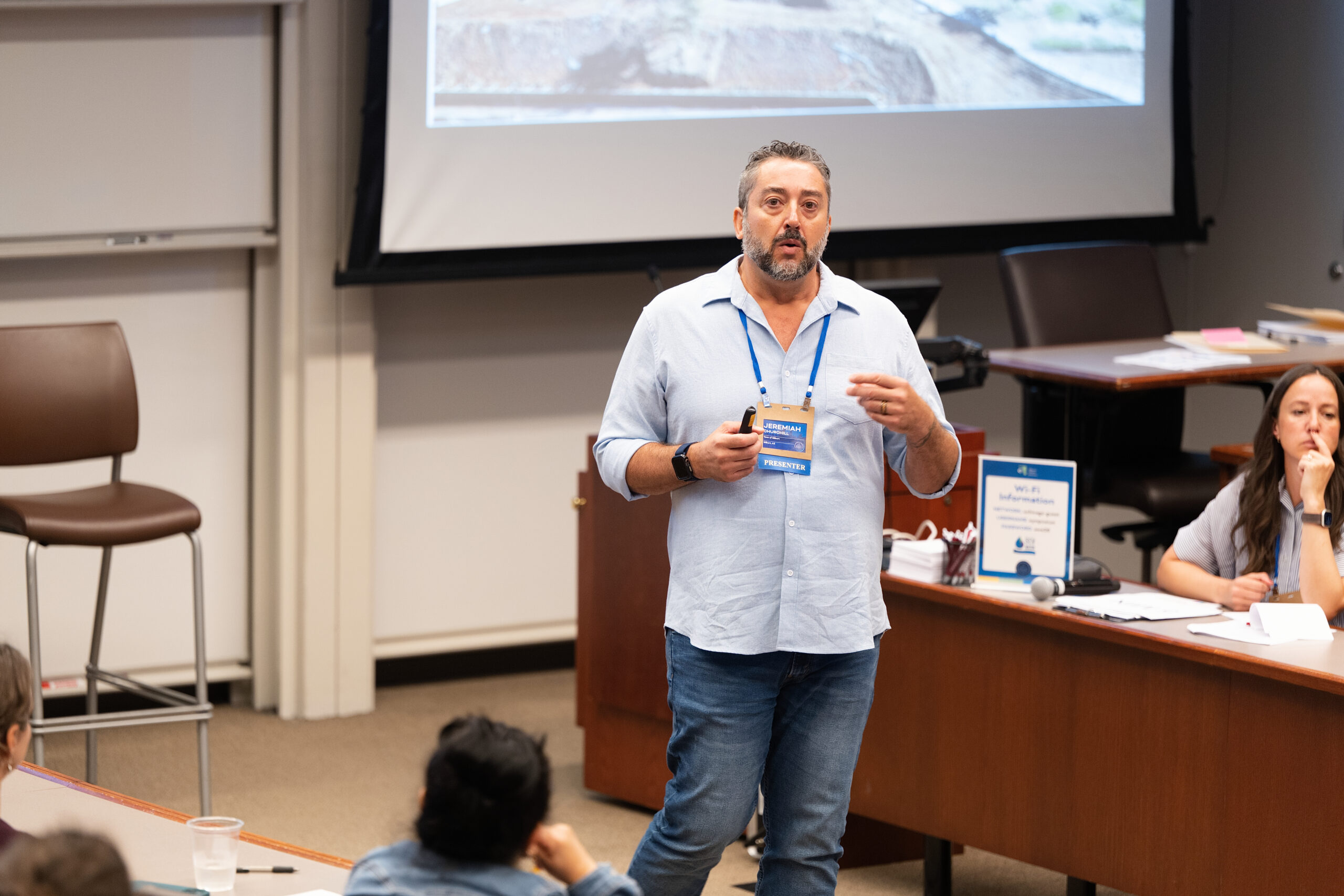 Male presenter in a light blue shirt with a blue lanyard and badge speaking to an audience in a seminar room, projector screen behind him.