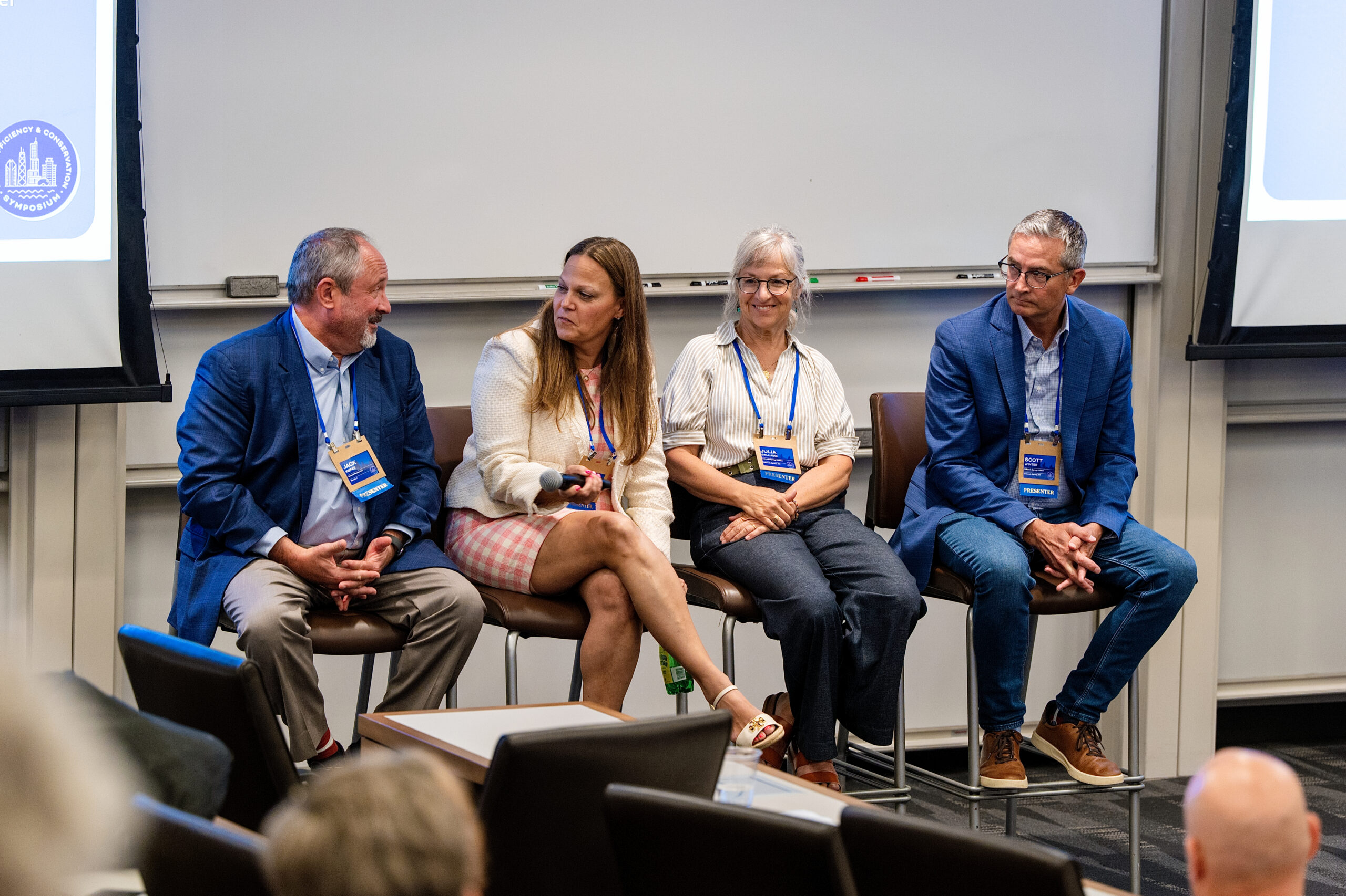 Panel of four professionals seated in a row on stage with name badges, participating in a conference discussion.