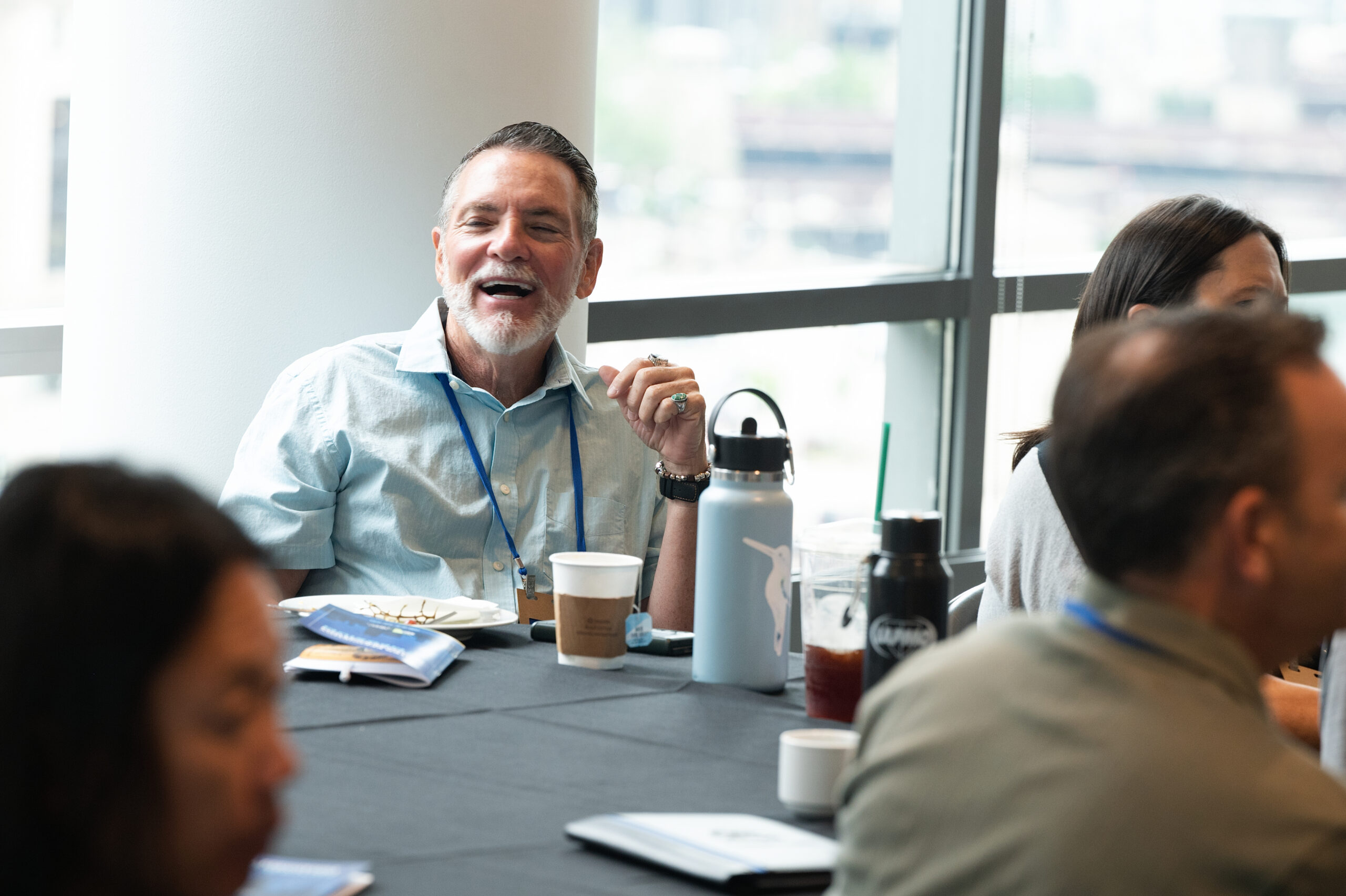 Middle-aged man with gray beard laughing at a table during a meeting, with cups and a water bottle nearby in a bright room.
