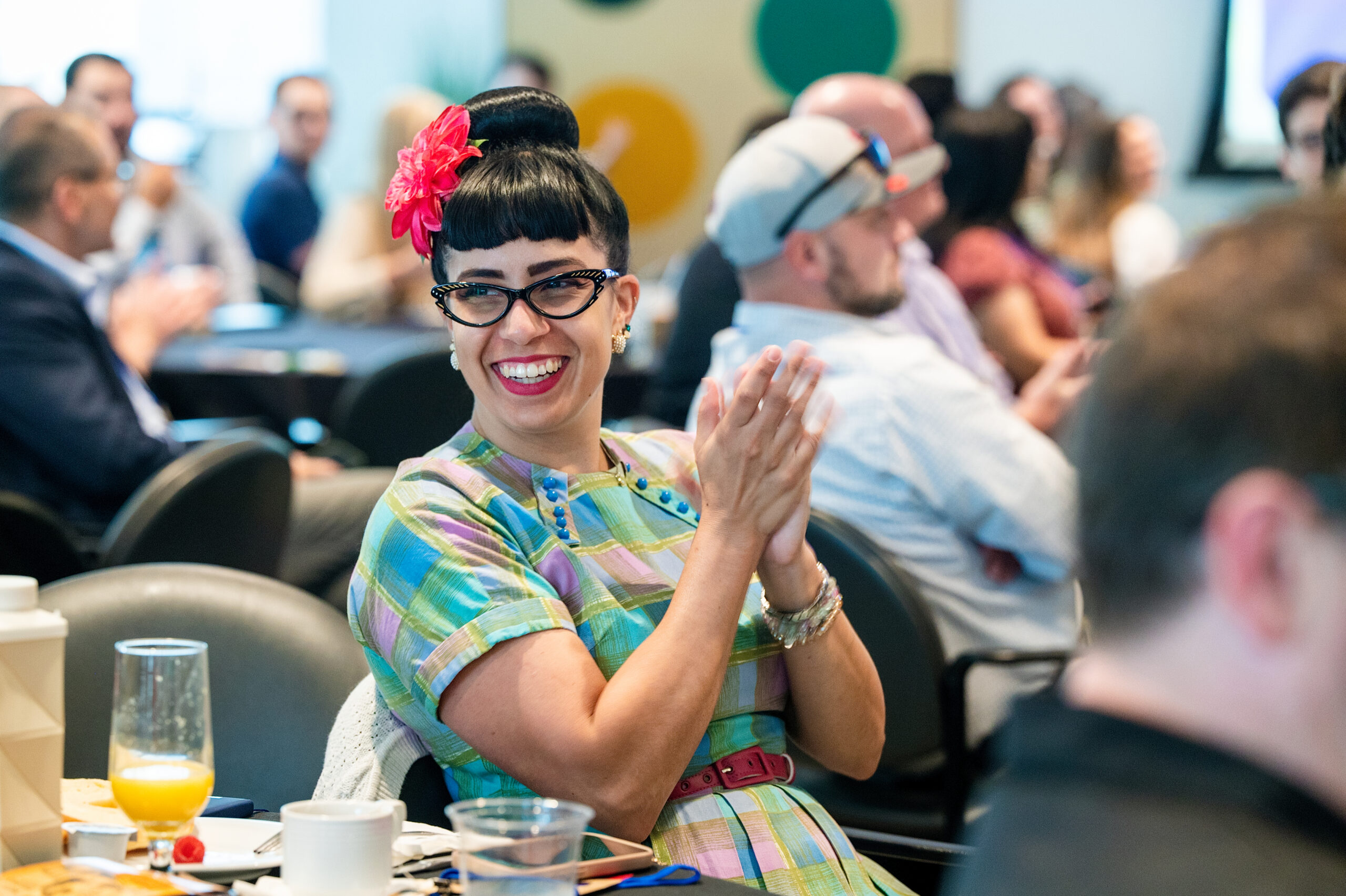 Smiling woman with a pink flower in her hair and black bangs clapping at a lively, crowded event.