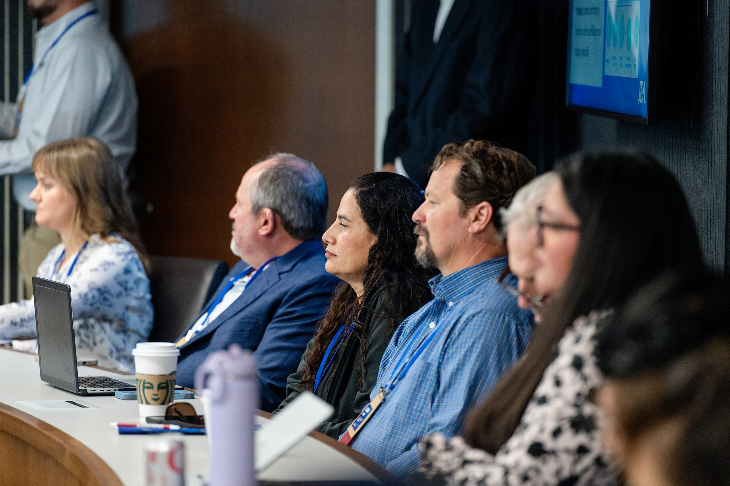 Group of professionals seated at a conference table, facing forward with laptops and coffee cups nearby.