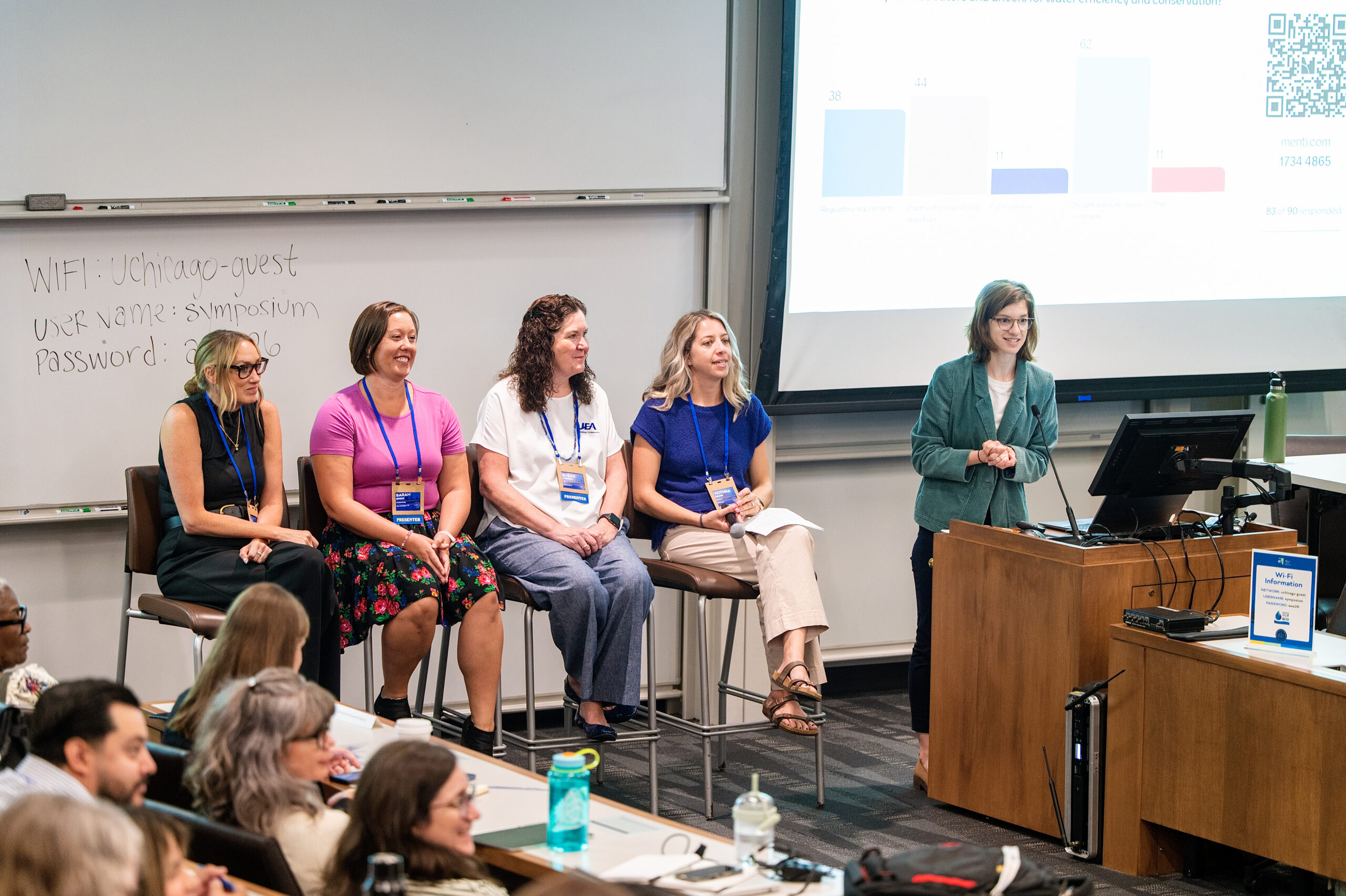 Five women panelists seated on stage in a classroom, with a presenter at a podium and a projected slide on a screen behind them.