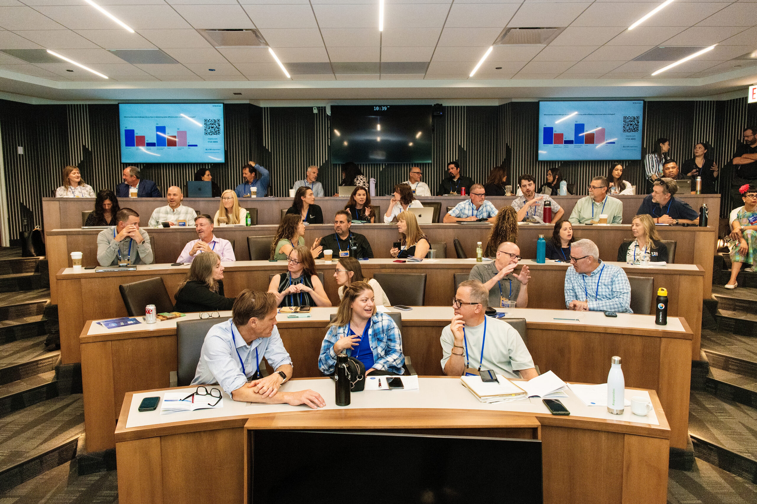 Large conference room filled with professionals seated at tiered desks, back screens showing charts and graphs.