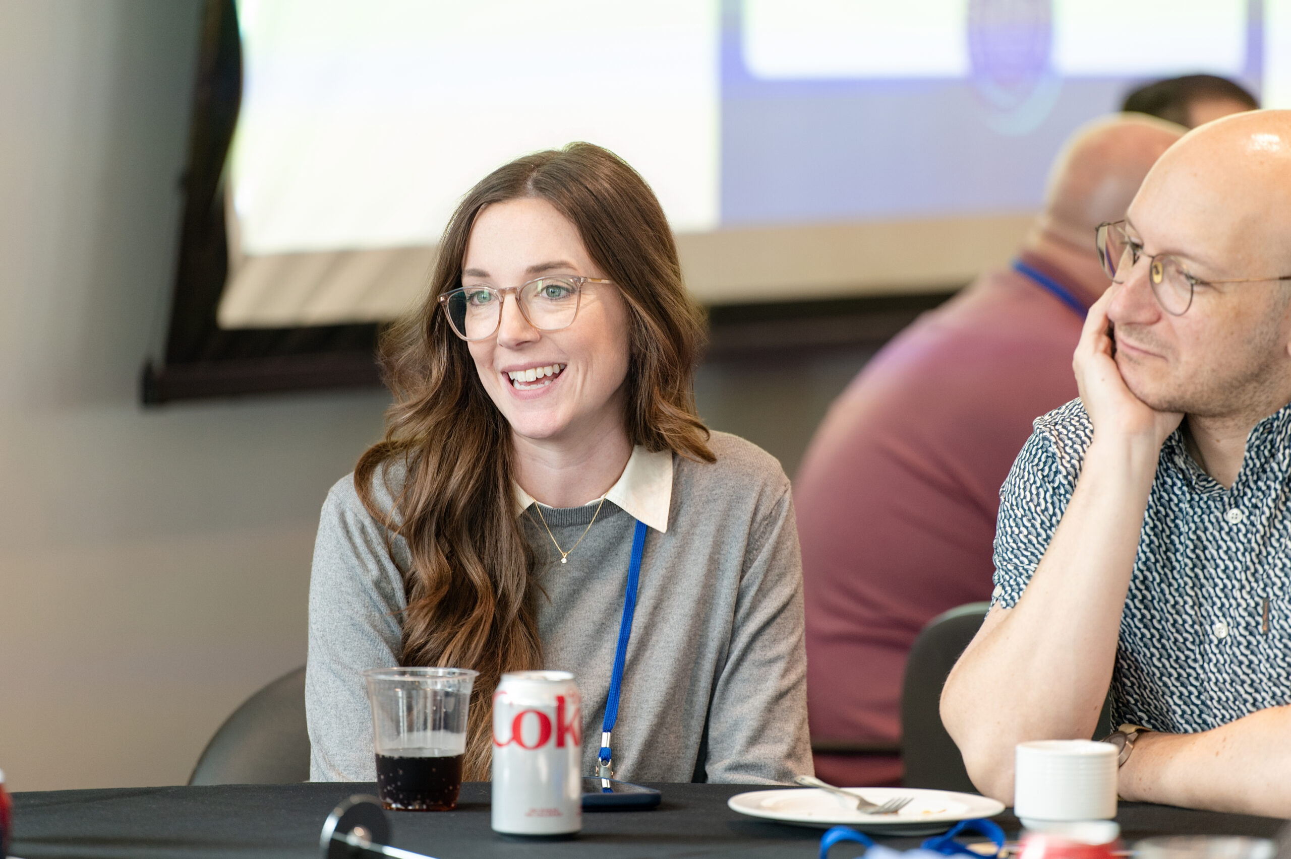 Smiling woman with glasses sits at a table next to a man resting his chin on his hand during a meeting, with soda cans in front of them.