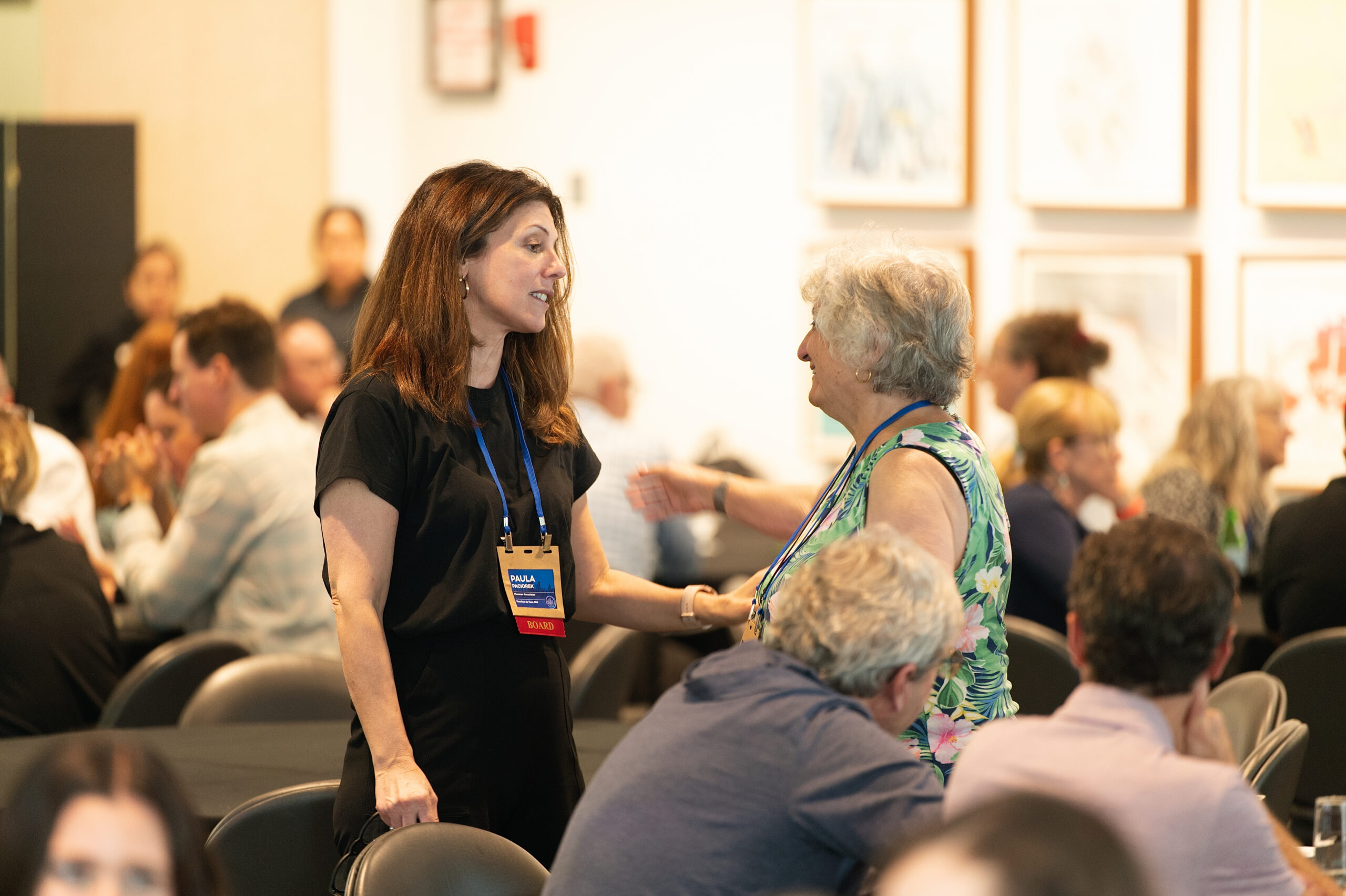 Two women stand and chat at a conference, one in a black outfit with a blue lanyard and name badge, the other in a floral top, as seated attendees blur in the background.