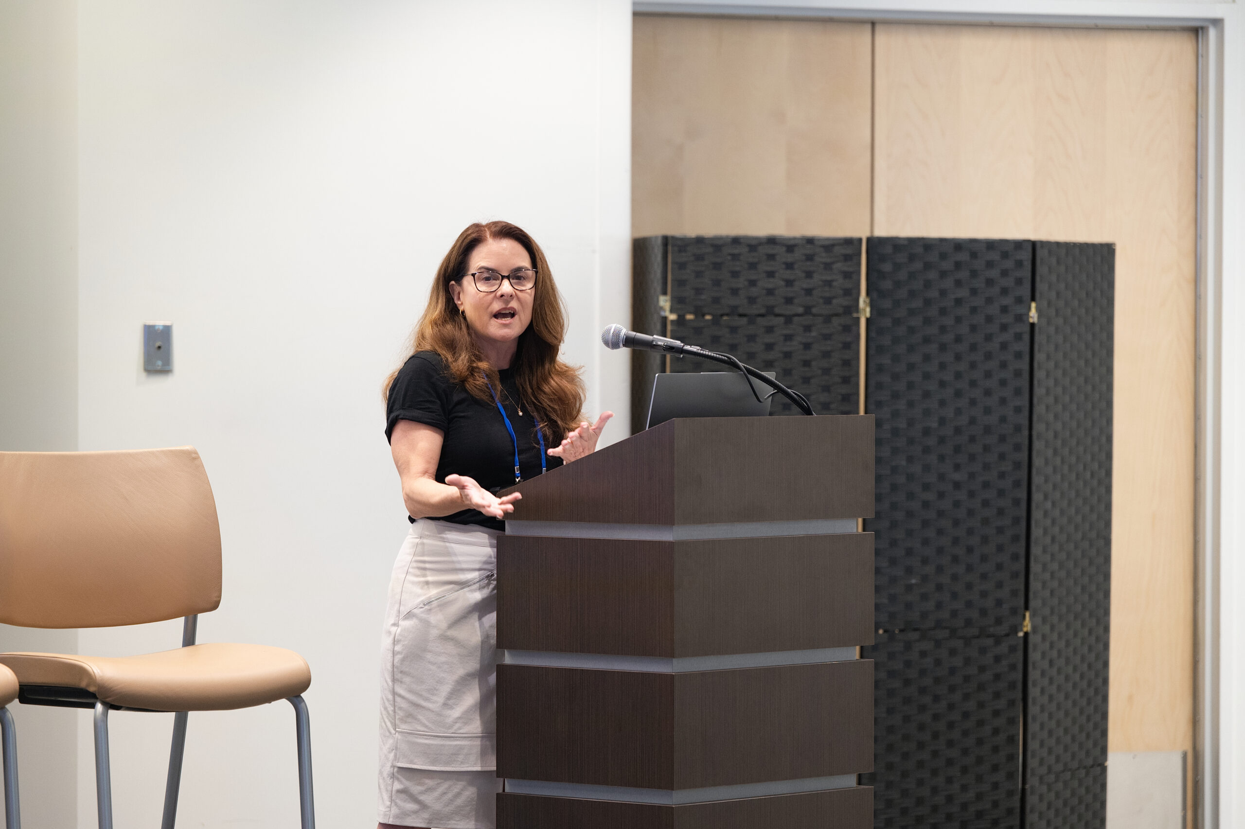 Woman speaking at a wooden podium with a microphone in a conference room; she wears a black top, light skirt, and glasses, gesturing with her hands as she addresses an audience.