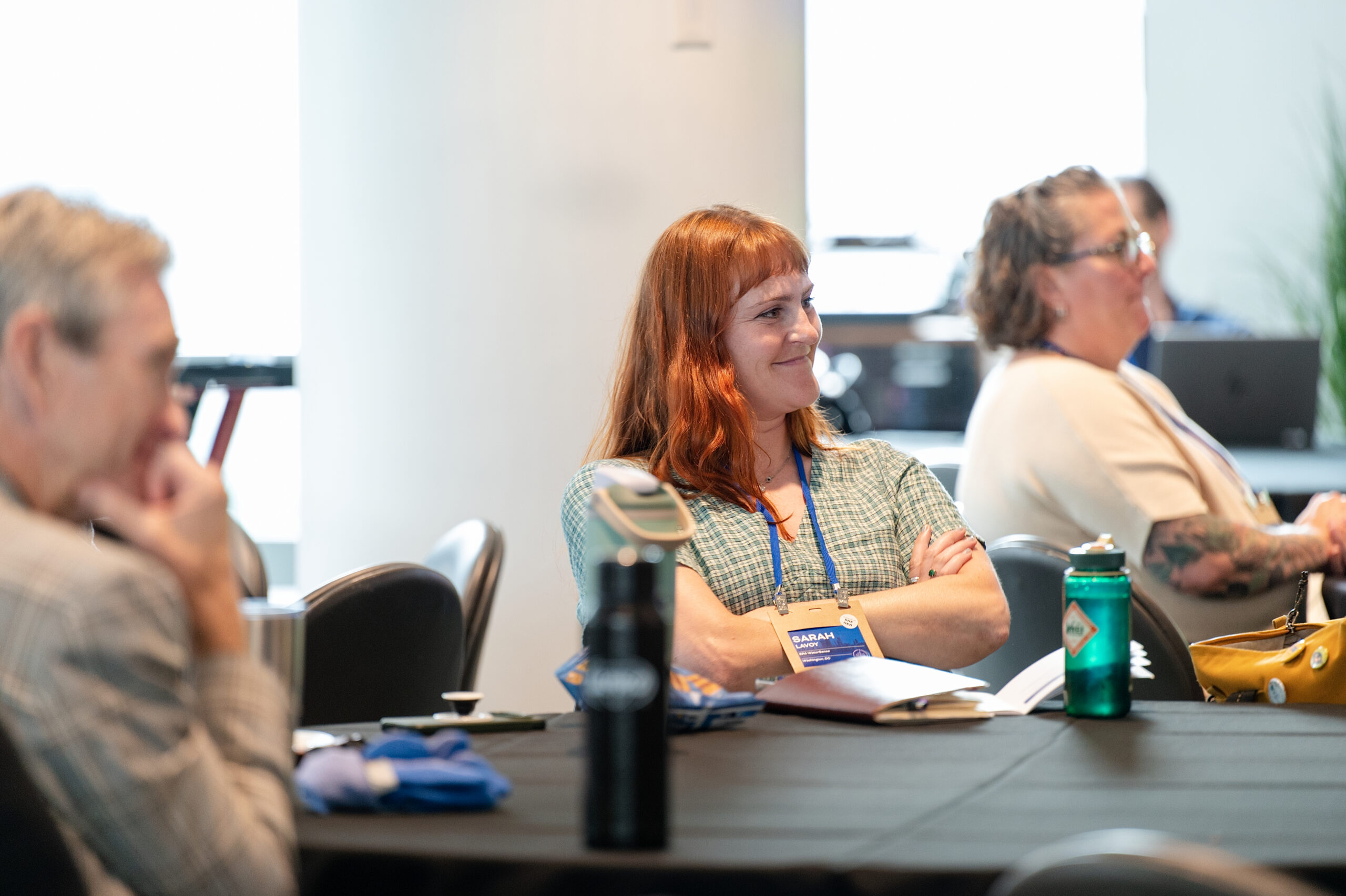 Woman with red hair and a name badge smiles at a conference table during a meeting.
