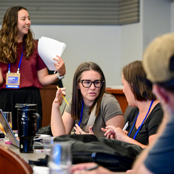 Group of coworkers around a conference table; a woman standing with papers leads the discussion.