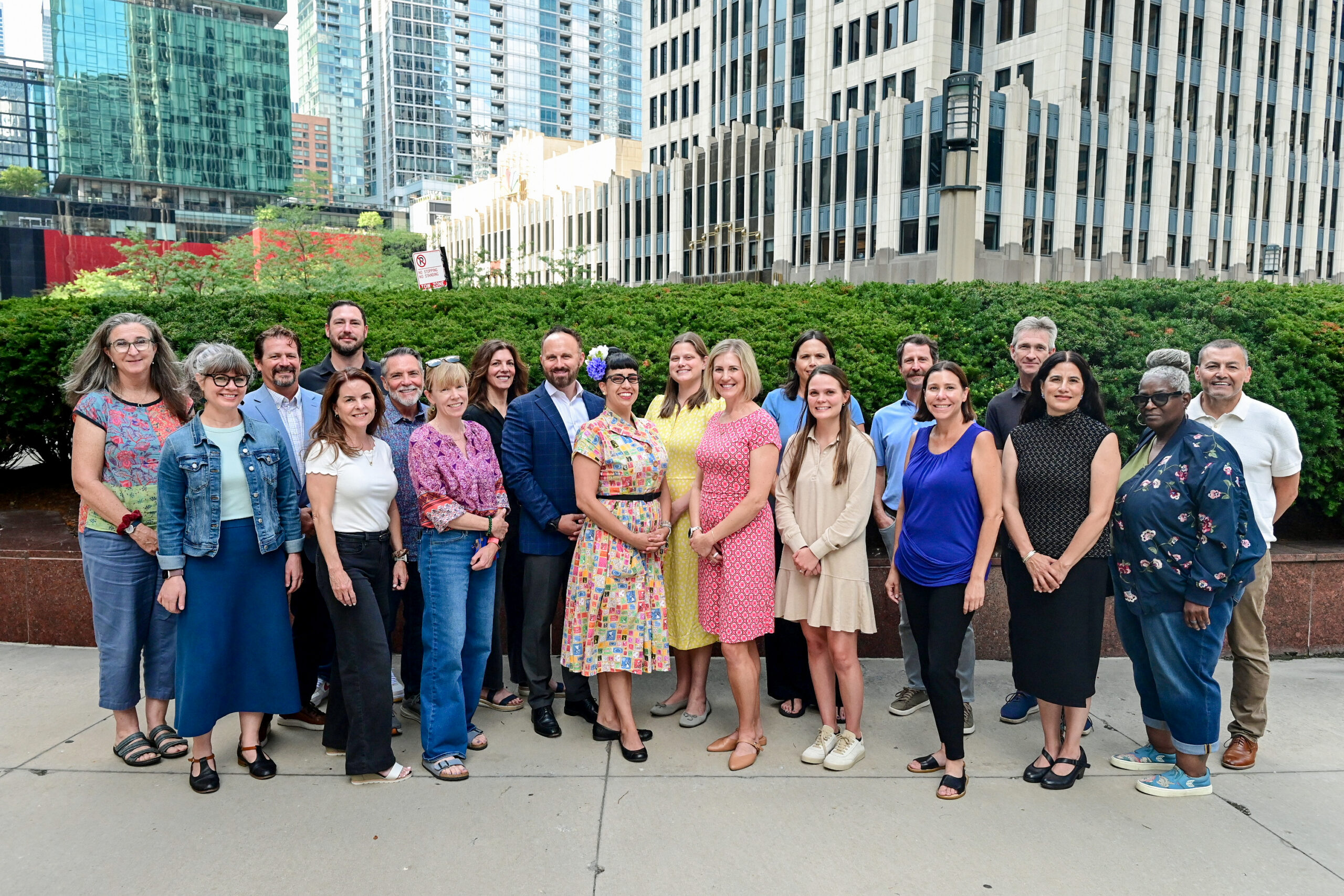Group of about 20 diverse adults posing together outdoors in front of modern glass and concrete office buildings.