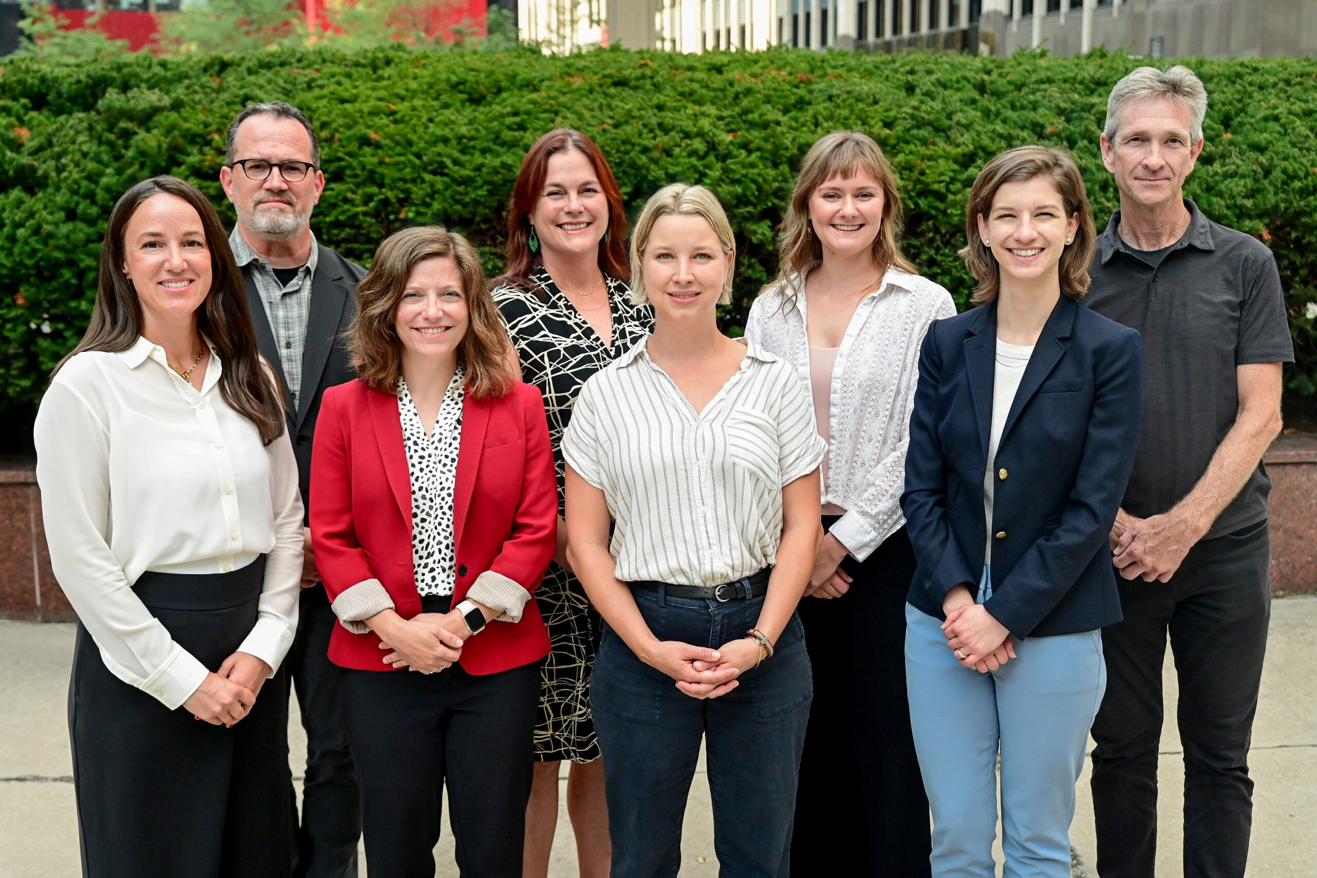 Eight adults posing outdoors in front of a green hedge, smiling at the camera.