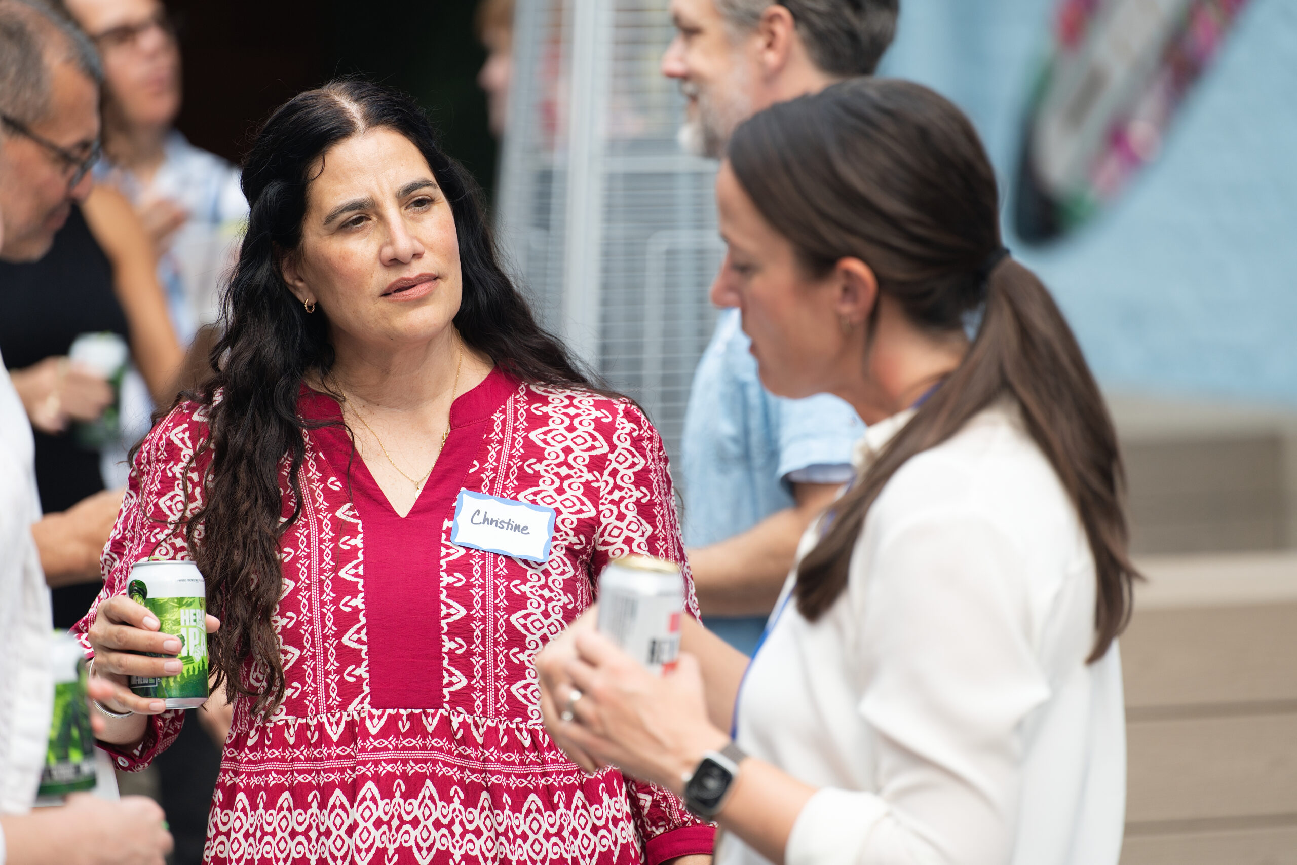 Woman in a red patterned dress with a name tag (Christine) converses with others at a social gathering, holding a beverage can.