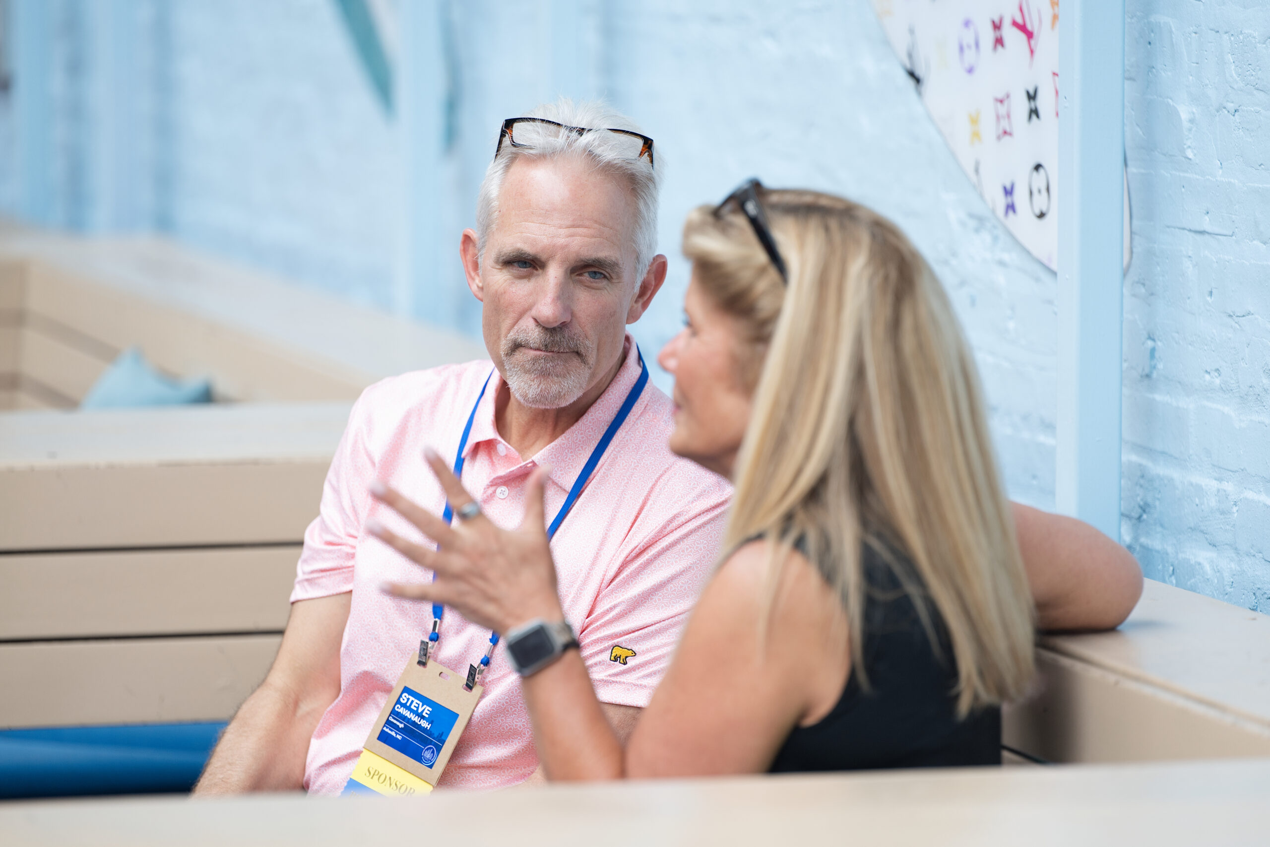 Two people conversing at a table outdoors; man in a pink shirt with sunglasses on his head and a visible event badge nearby.