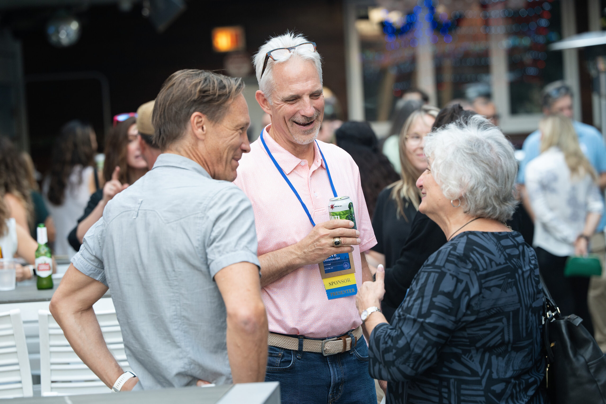 Adults mingle at an outdoor event; center man in a pink shirt with a sponsor badge smiles while talking with others.