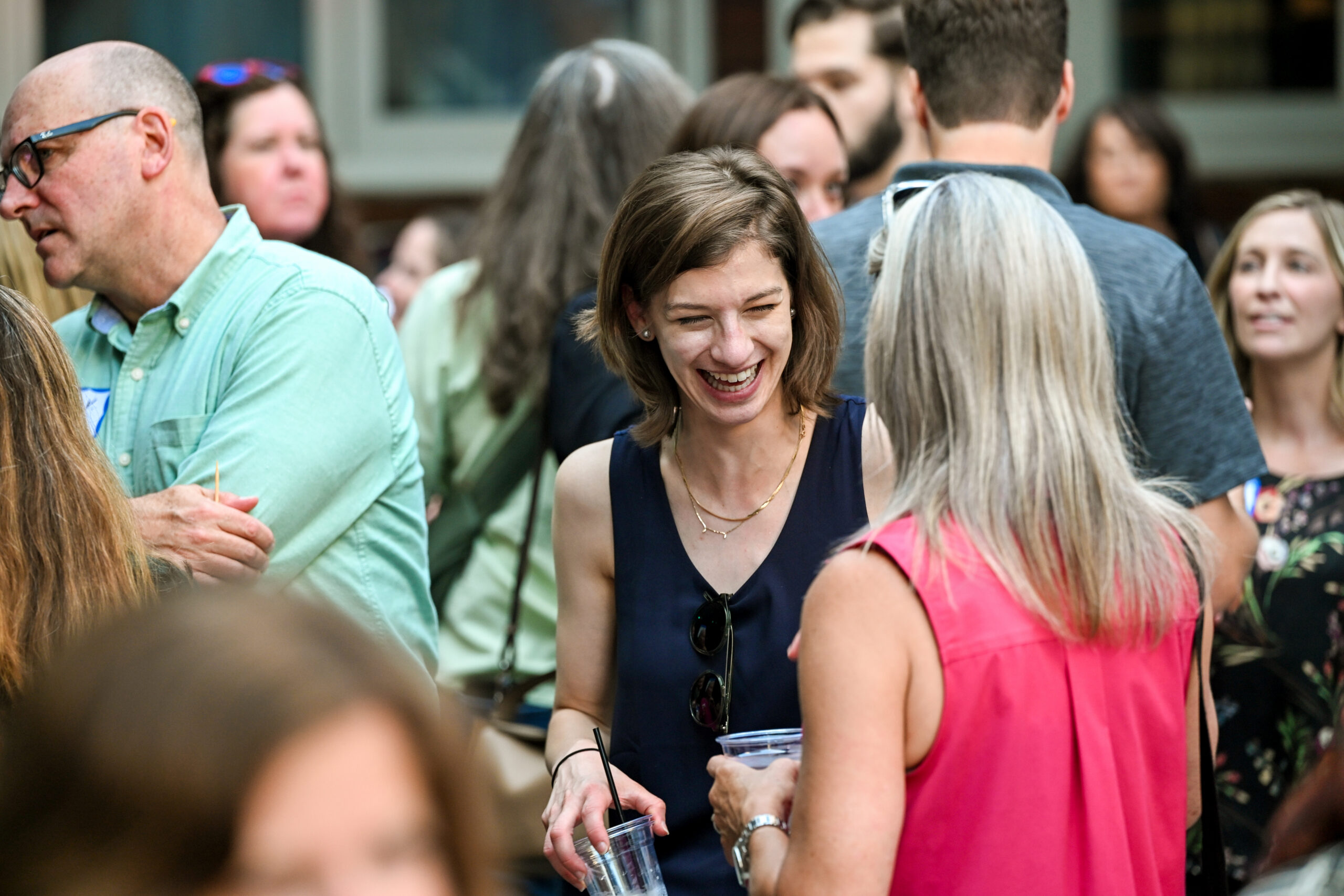 Smiling woman in a navy dress holding a cup with a straw at a crowded outdoor party.
