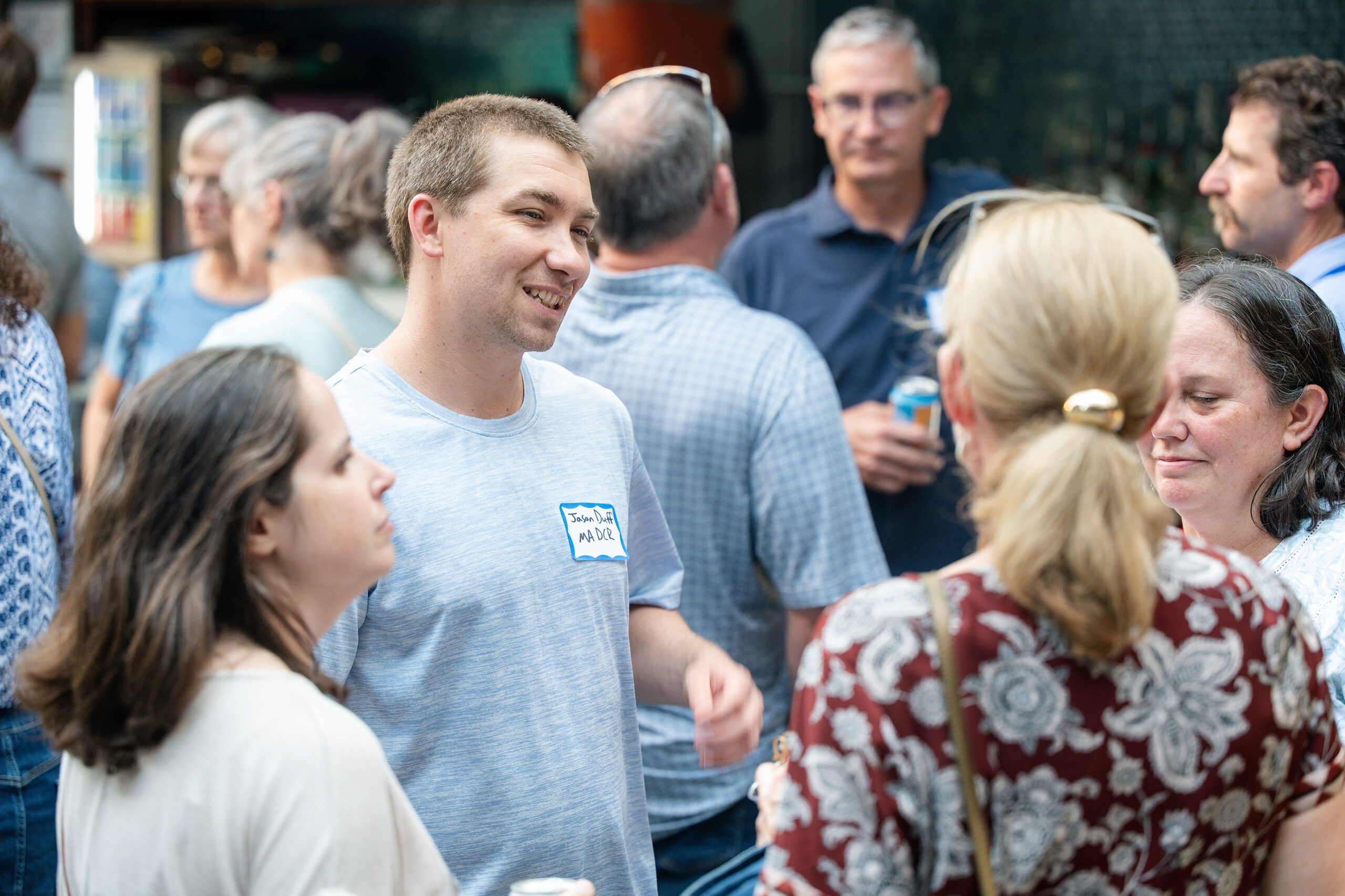 Smiling man in a light blue T-shirt with a name tag talking to a crowd at an outdoor gathering.