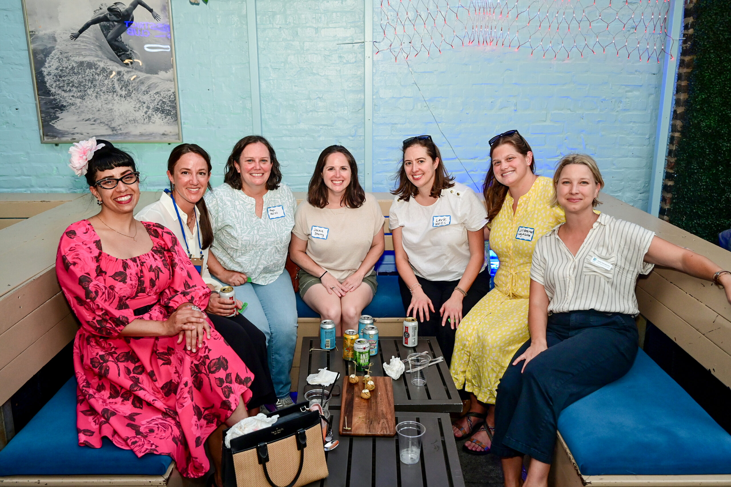 Group of seven women sitting together on a bench-style seating area, smiling at a casual gathering with drinks and snacks on a table in front of them.