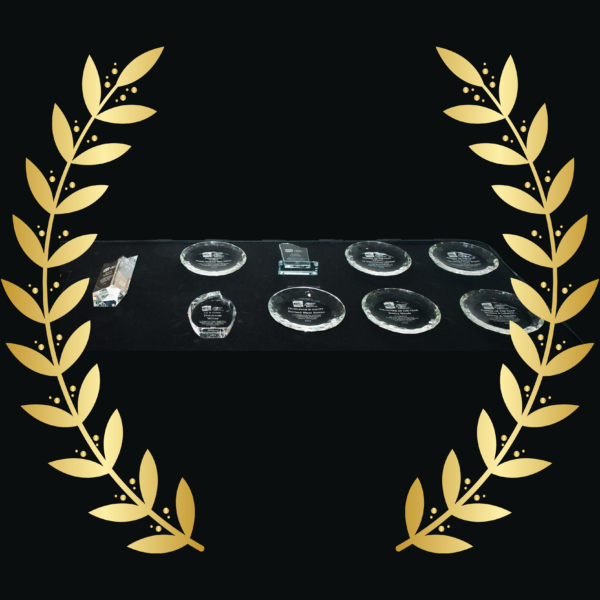 Stage backdrop featuring gold laurel wreaths on a black background with glass award plaques arranged on a table in front.