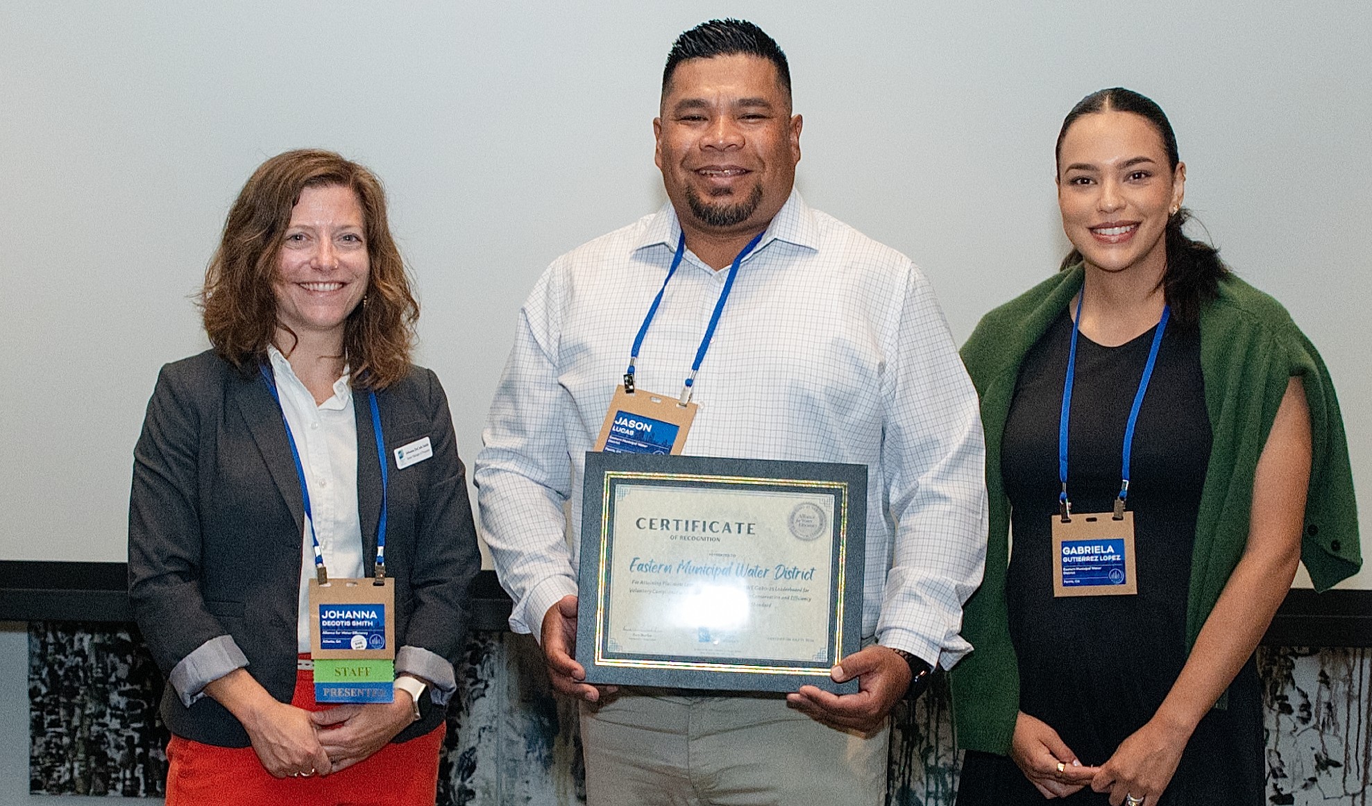 Three colleagues pose with a framed certificate of recognition awarded to the Eastern Municipal Water District, center man smiling holding the certificate, two women on sides wearing badges.